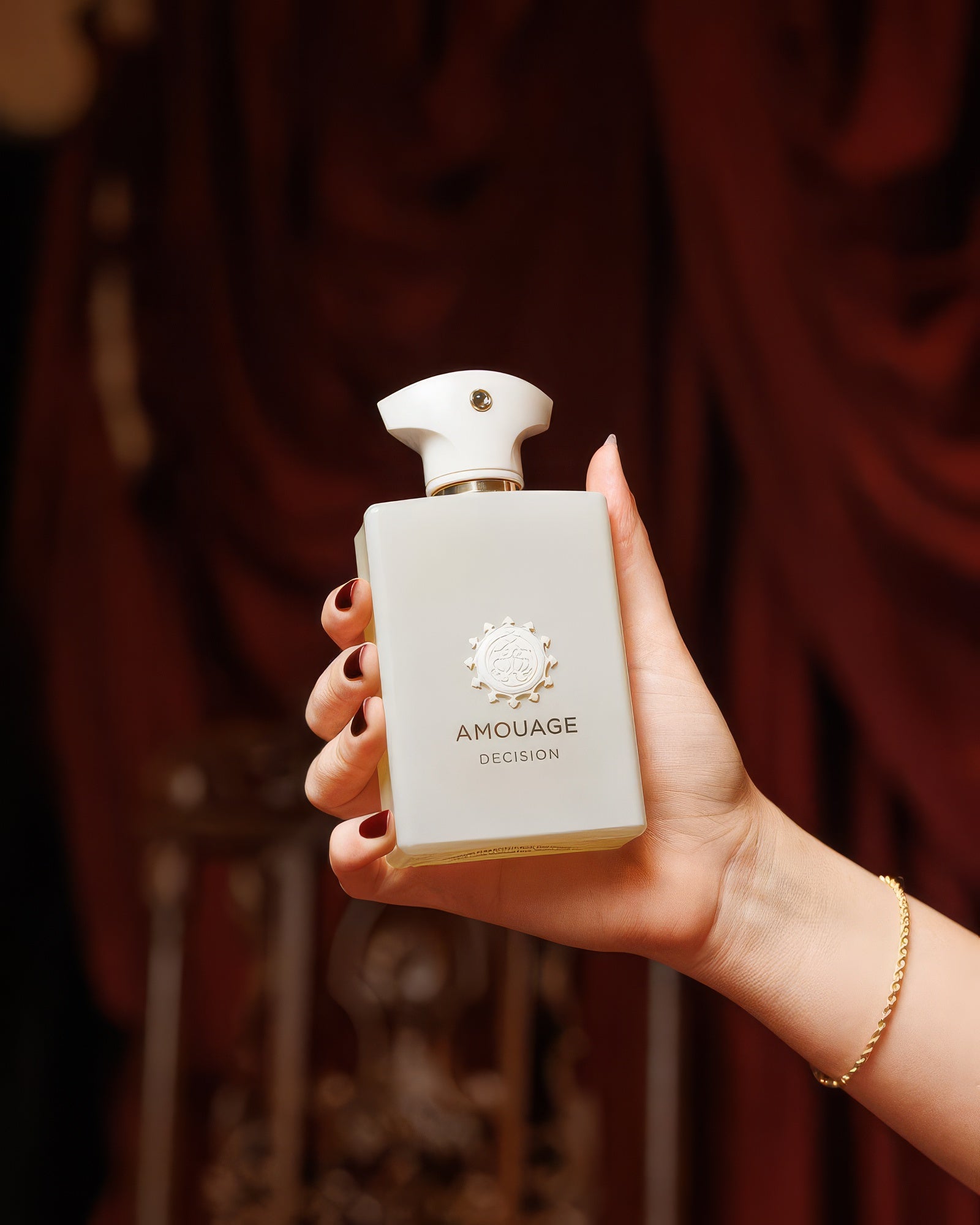 Amouage Decision محمولة بيد مُعتنى بها أمام ستائر مخملية حمراء داكنة، عينة عطر نيش بخور مدخّن