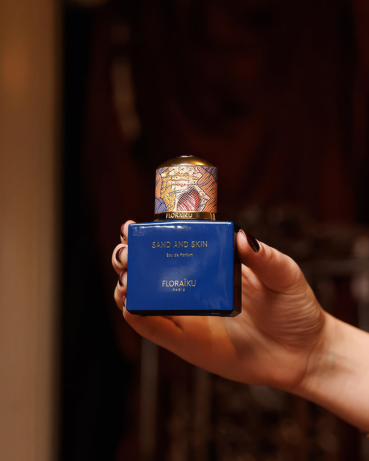 Hand hält den blauen Floraiku Sand and Skin Flakon in warmem, gedämpftem Licht, pudrig-moschusartige Duftprobe