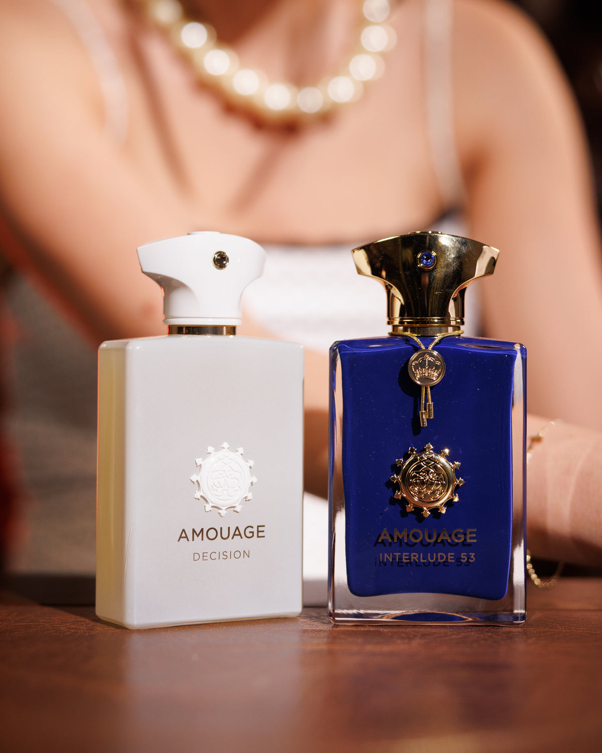 Parlak bir bar tezgâhında beyaz Amouage Decision şişesinin yanında altın kapaklı, taç süslemeli mavi Amouage Interlude 53 şişesi, arkada bulanık bir model, dumanlı tütsü notalı niş parfüm dekantı