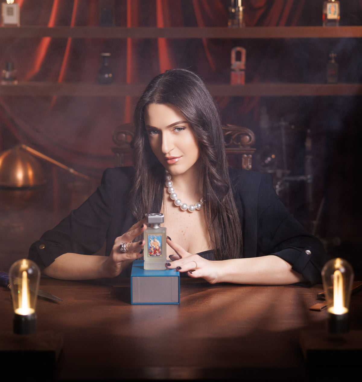 Argos Neptune's Trident on its blue box, framed by the hands of a woman in a black blazer and pearls at a wooden desk, Edison lamps aglow on each side, shelves in haze behind