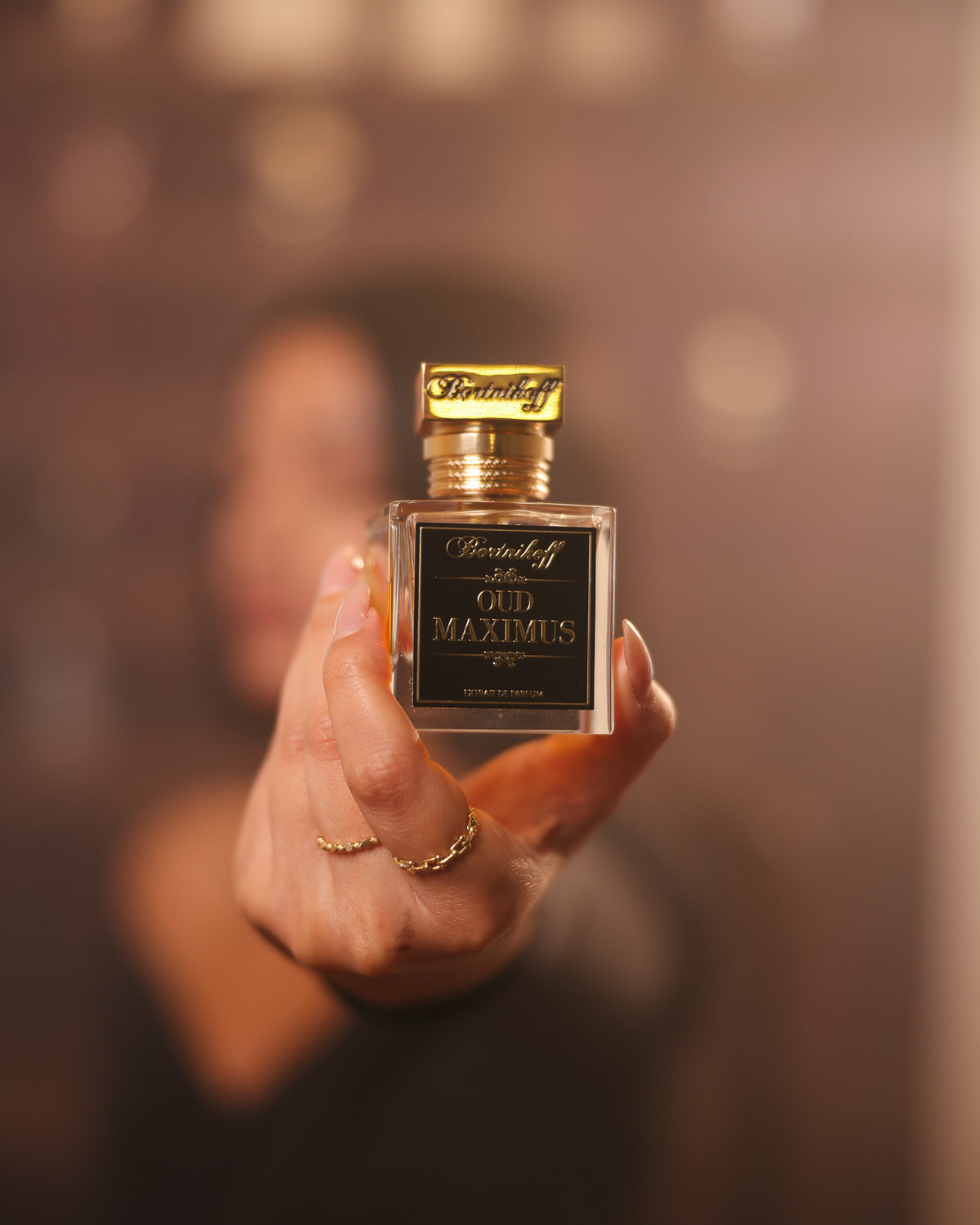 Bortnikoff Oud Maximus, flacone quadrato porto verso l'obiettivo da una mano con anelli d'oro, bokeh caldo, decant extrait de parfum oud animalico