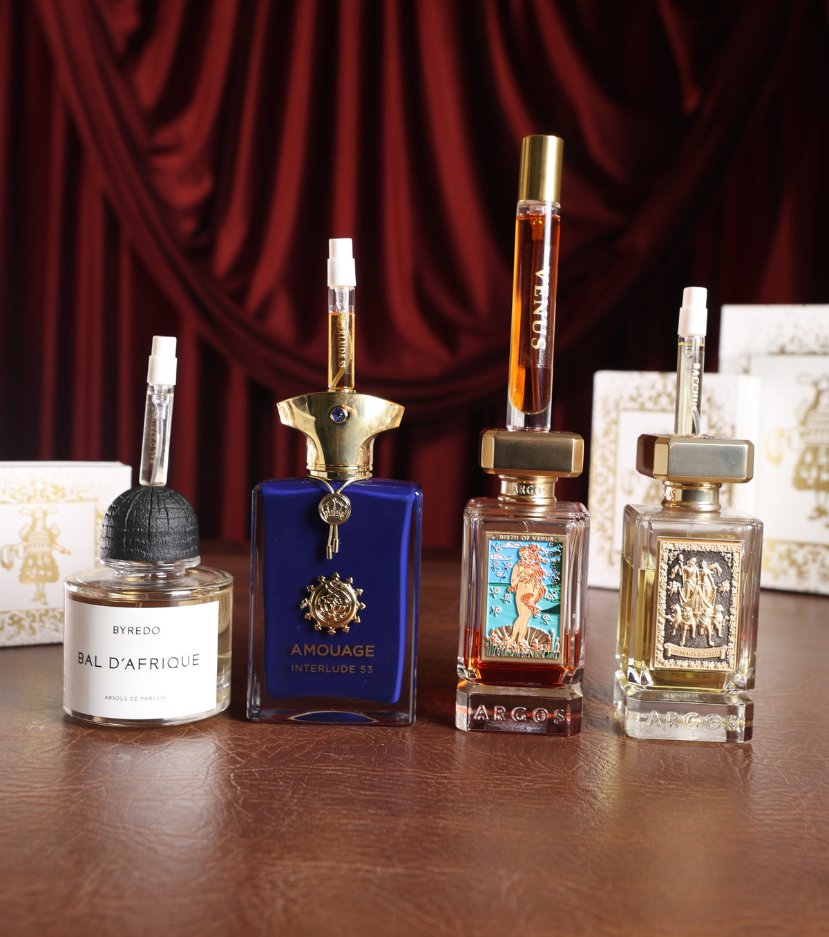 Byredo Bal D'Afrique Absolu, Amouage Interlude 53, Argos Birth of Venus and Argos Triumph of Bacchus, each topped with a Gouttes Rares decant, gold-foil boxes and red velvet behind