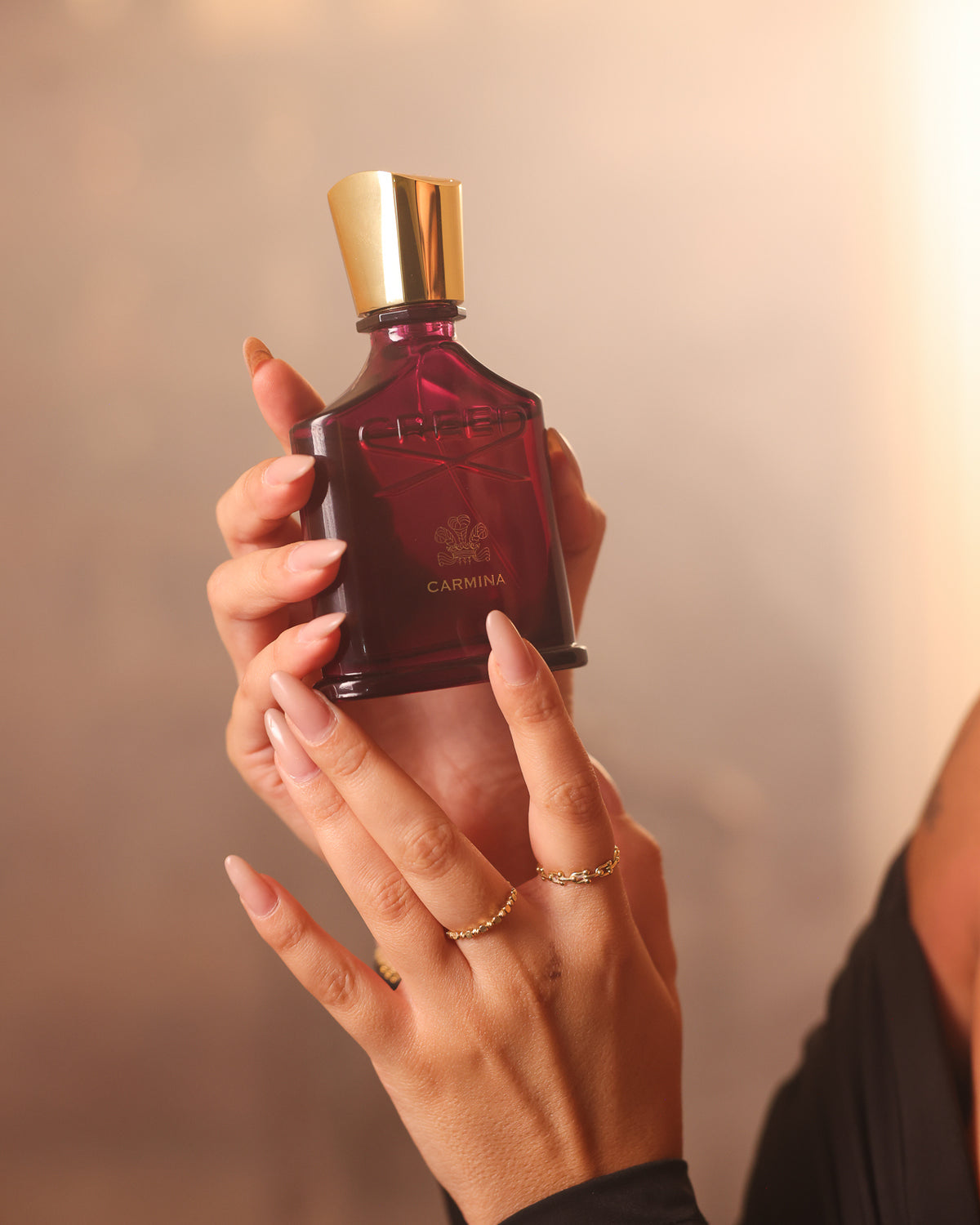 Creed Carmina burgundy bottle with gold cap held upright in two hands with long nude nails and gold rings, warm blurred backdrop, fruity floral niche perfume decant