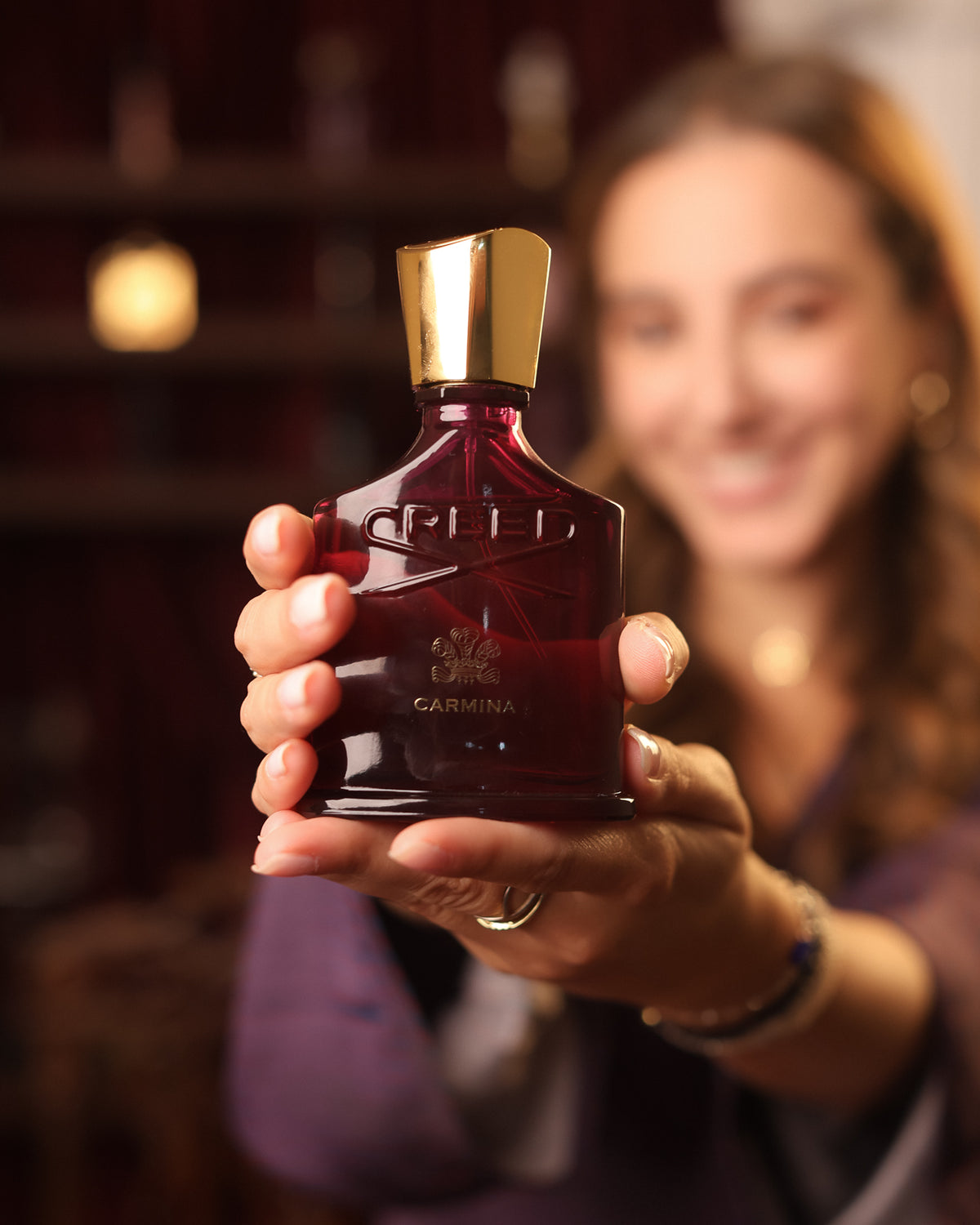 Creed Carmina burgundy bottle with gold cap held out to the camera by a smiling woman in a purple top, warm blur, fruity floral niche perfume decant