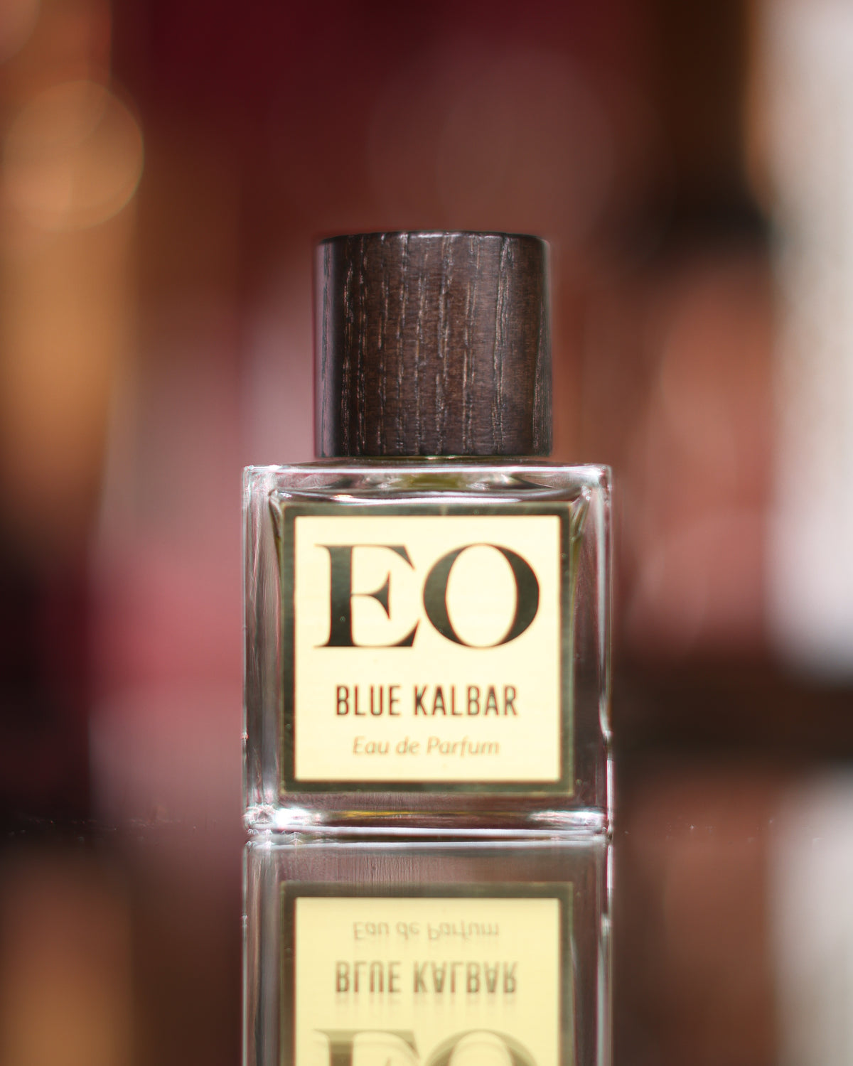 Ensar Oud Blue Kalbar square bottle with dark wood cap and cream EO label reflected in a dark mirror before warm bokeh, oud eau de parfum decant