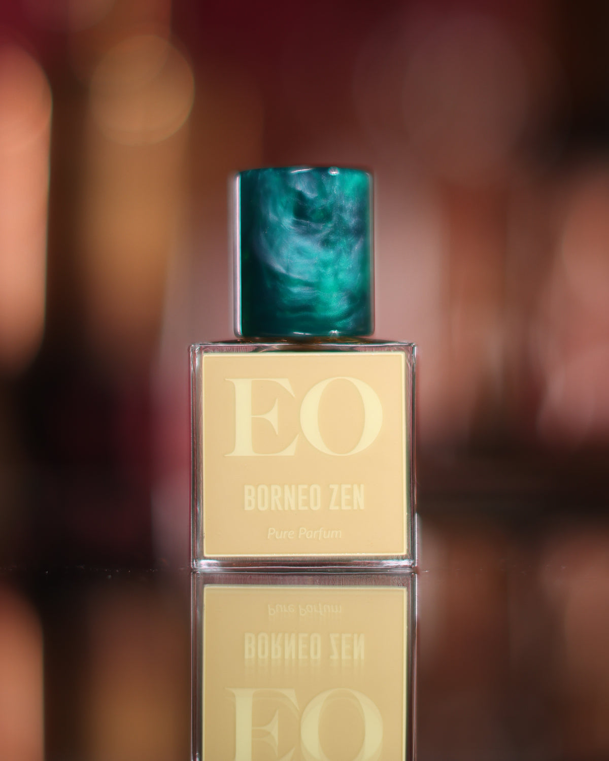 Ensar Oud Borneo Zen: Trifecta square bottle, marbled teal cap and pale gold EO label reflected in a dark mirror before warm bokeh, oud extrait de parfum decant