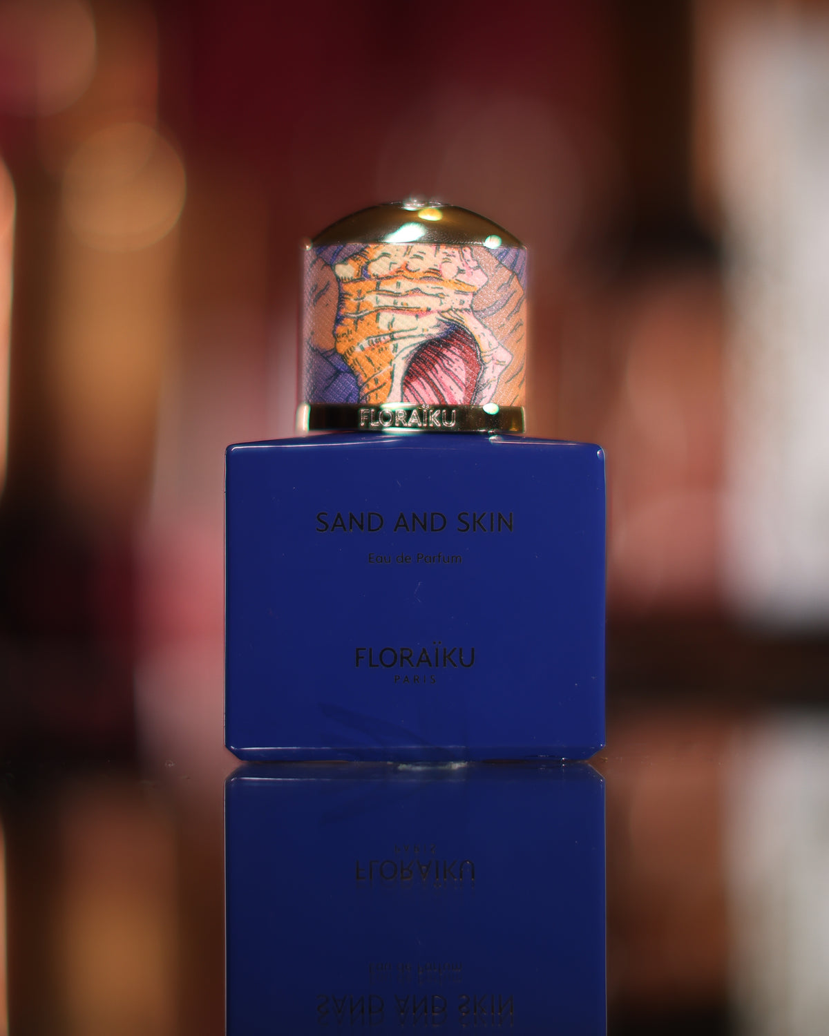 Kobaltblaue Floraïku Sand and Skin Flasche mit illustrierter Muschelkappe, gespiegelt auf einem dunklen Spiegel, warme unscharfe Lichter dahinter, Abfüllung eines moschusartigen Vanille-Nischenparfums