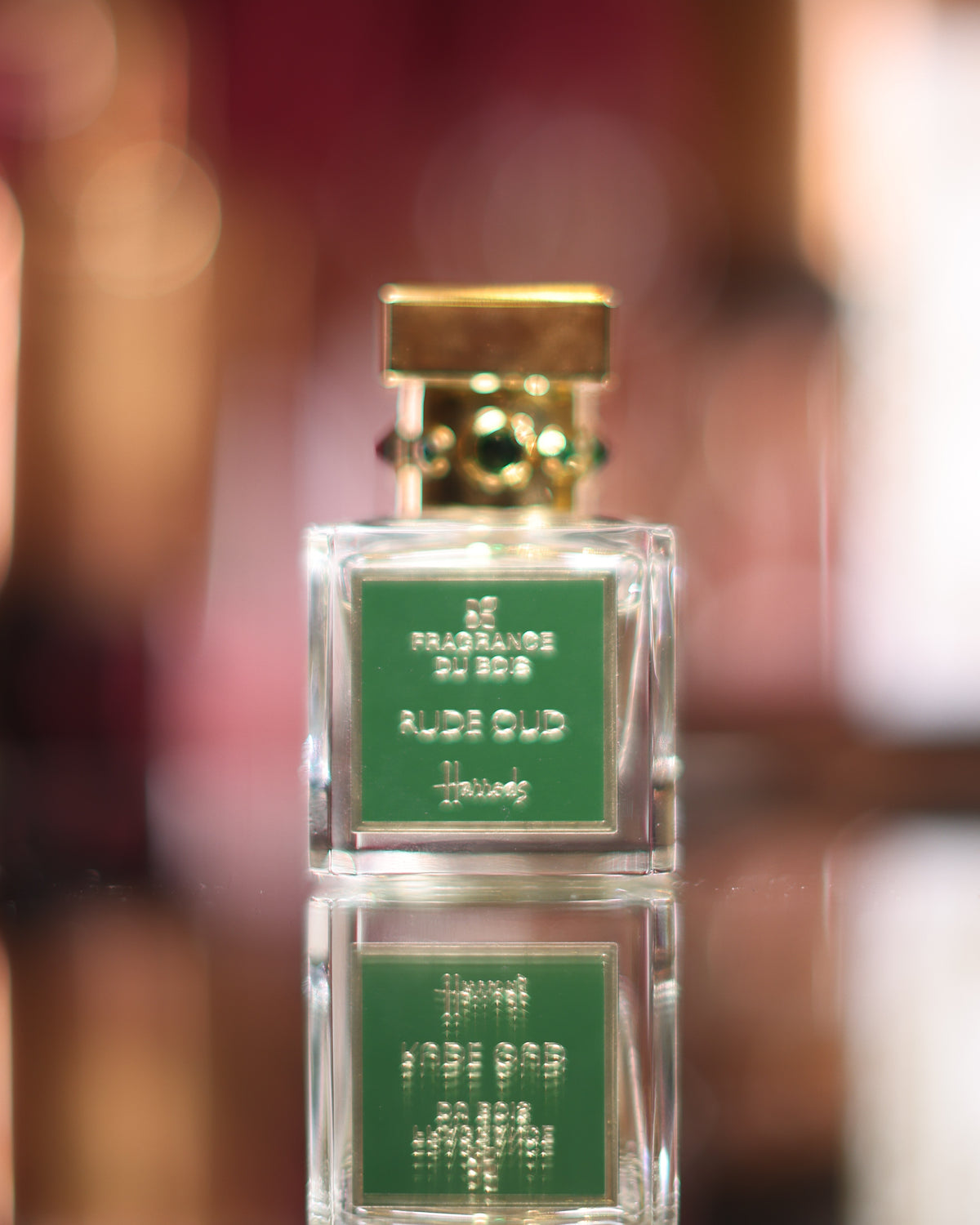 Fragrance Du Bois Rude Oud emerald label flacon with gold cap set with green stones reflected on a dark mirror, warm blurred lights behind, oud extrait de parfum decant
