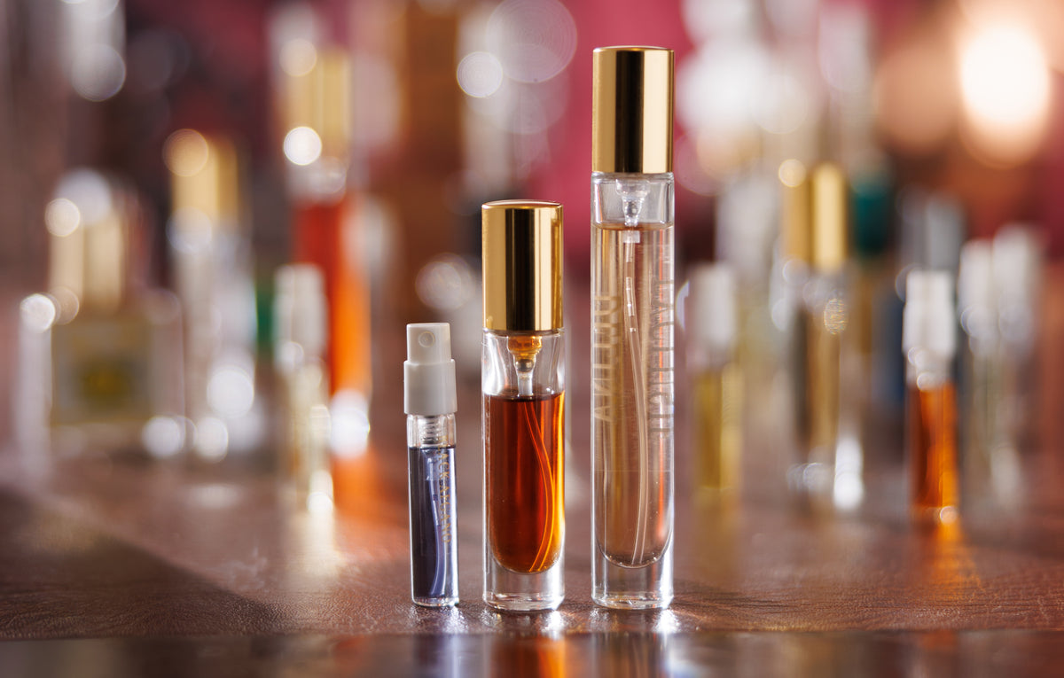 عبوات Gouttes Rares المقسّمة بثلاثة أحجام، بخّاخ عيّنة بسعة 2ml وبخّاخان بسعة 5ml و10ml بغطاءين ذهبيين، قائمة على مكتب لامع أمام ضبابية دافئة من زجاجات النيش