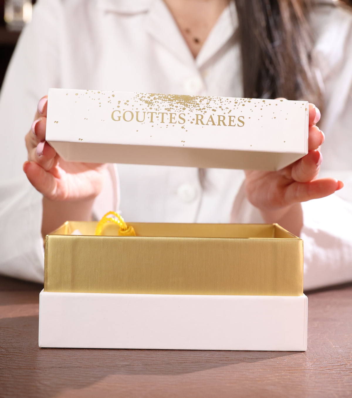 Hands lifting the white Gouttes Rares lid with gold lettering and gold speckles above its gold lined box, the pouch cord inside, niche perfume decant gift box