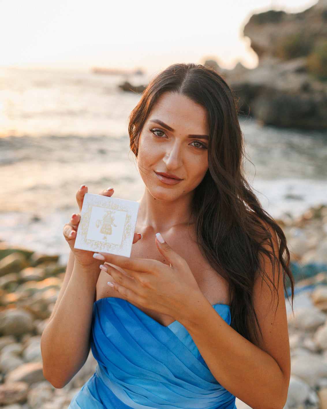 Gouttes Rares Niche Perfume Discovery Sets 4 x 2ml box held up by a woman in a blue strapless dress on a pebble beach at sunset, curated sampler box