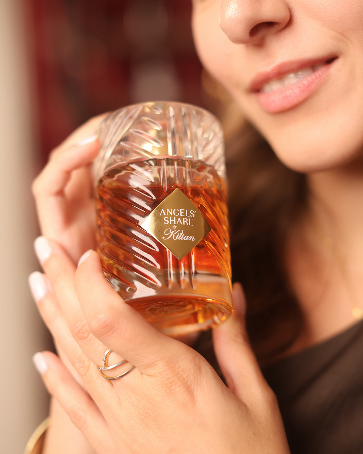 Kilian Paris Angels’ Share, frasco tallado como un cristal sostenido entre dedos de puntas blancas bajo los labios sonrientes de una mujer, decant de perfume nicho de coñac licoroso