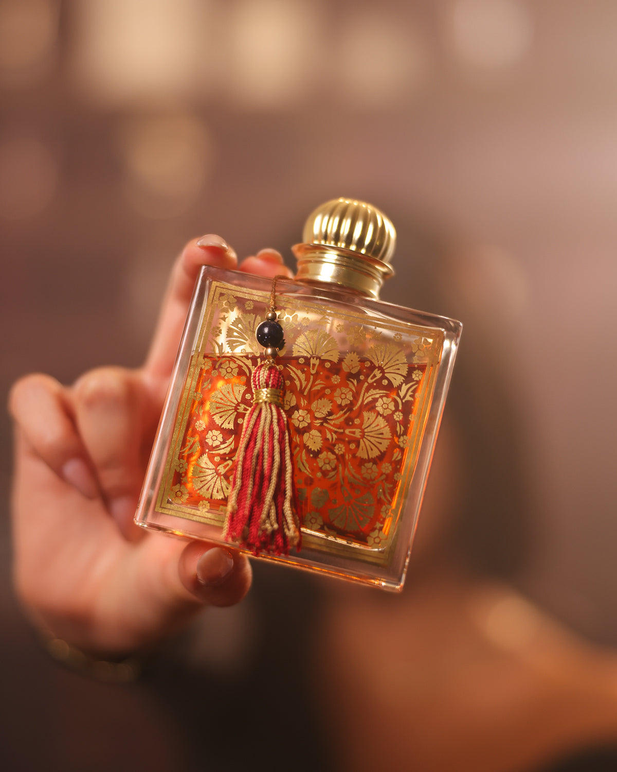 MDCI Chypre Palatin, frasco quadrado inclinado numa mão, tampa dourada canelada e borla vermelha reluzindo contra luzes quentes desfocadas, decant eau de parfum chipre