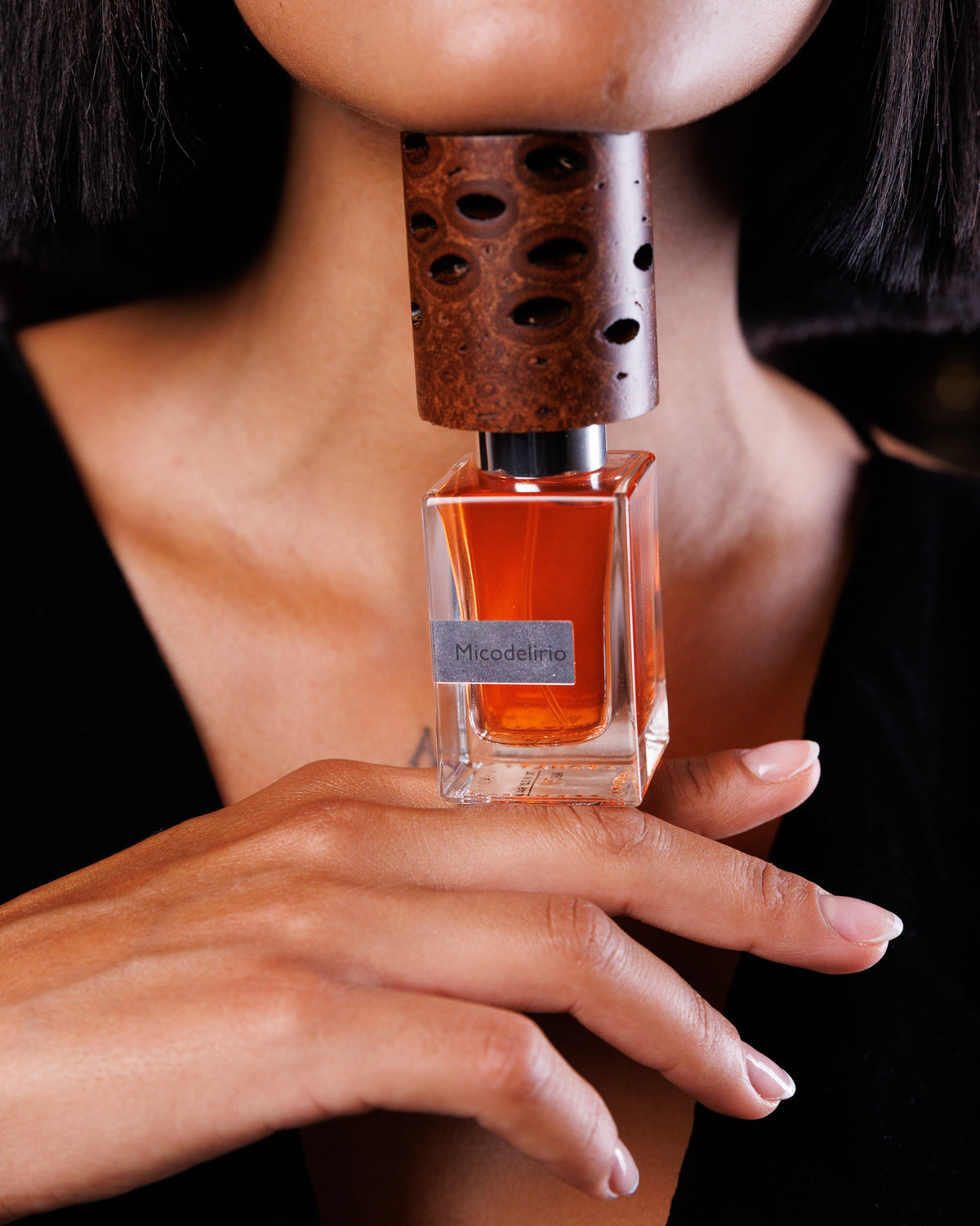 Flacone di Nasomatto Micodelirio, tappo a baccello di banksia accostato sotto il mento di una modella, il flacone in equilibrio sul dorso delle sue dita sopra velluto nero, decant di profumo di nicchia