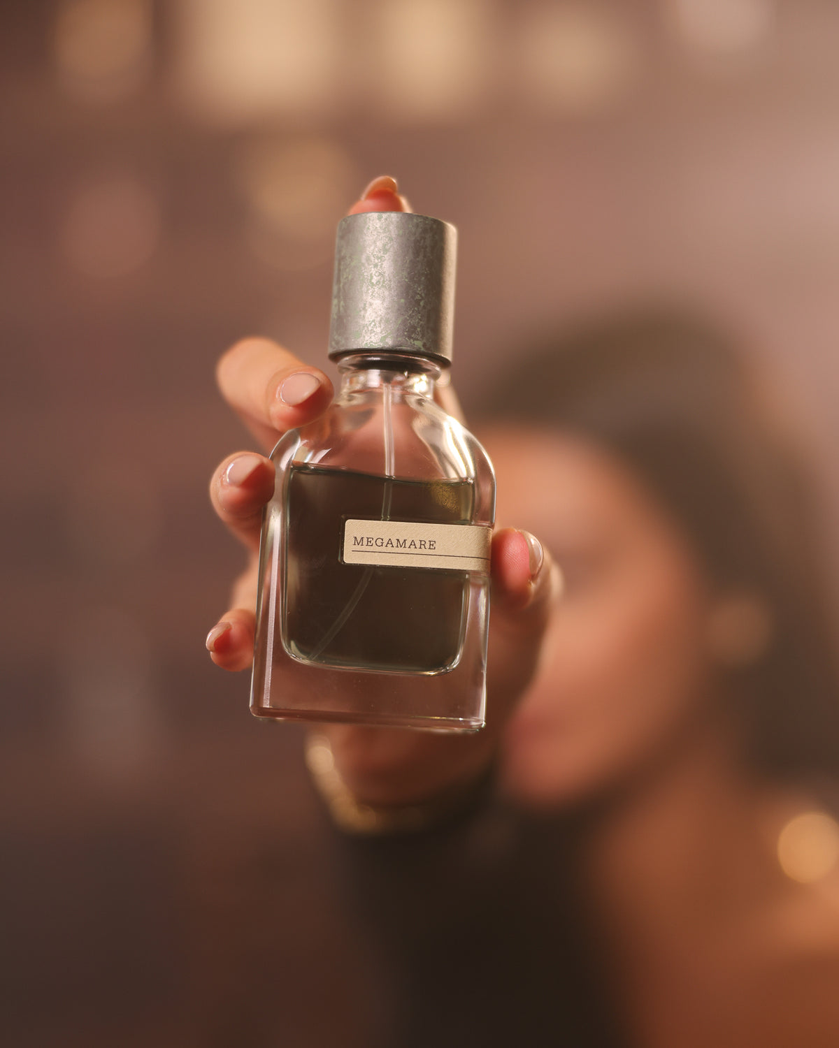 Orto Parisi Megamare, frasco con tapón de metal envejecido tendido hacia la cámara ante un rostro desenfocado y un bokeh cálido, decant de perfume nicho acuático