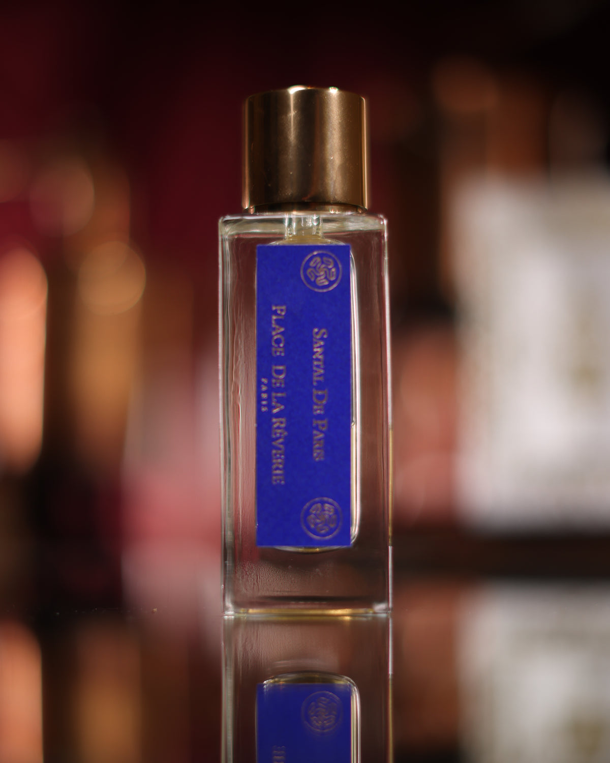Klare Place de la Rêverie Santal de Paris Flasche mit königsblauem Etikett und Goldkappe, gespiegelt auf einem schwarzen Spiegel vor unscharfen roten Lichtern, Abfüllung eines pudrigen Sandelholz-Extraits