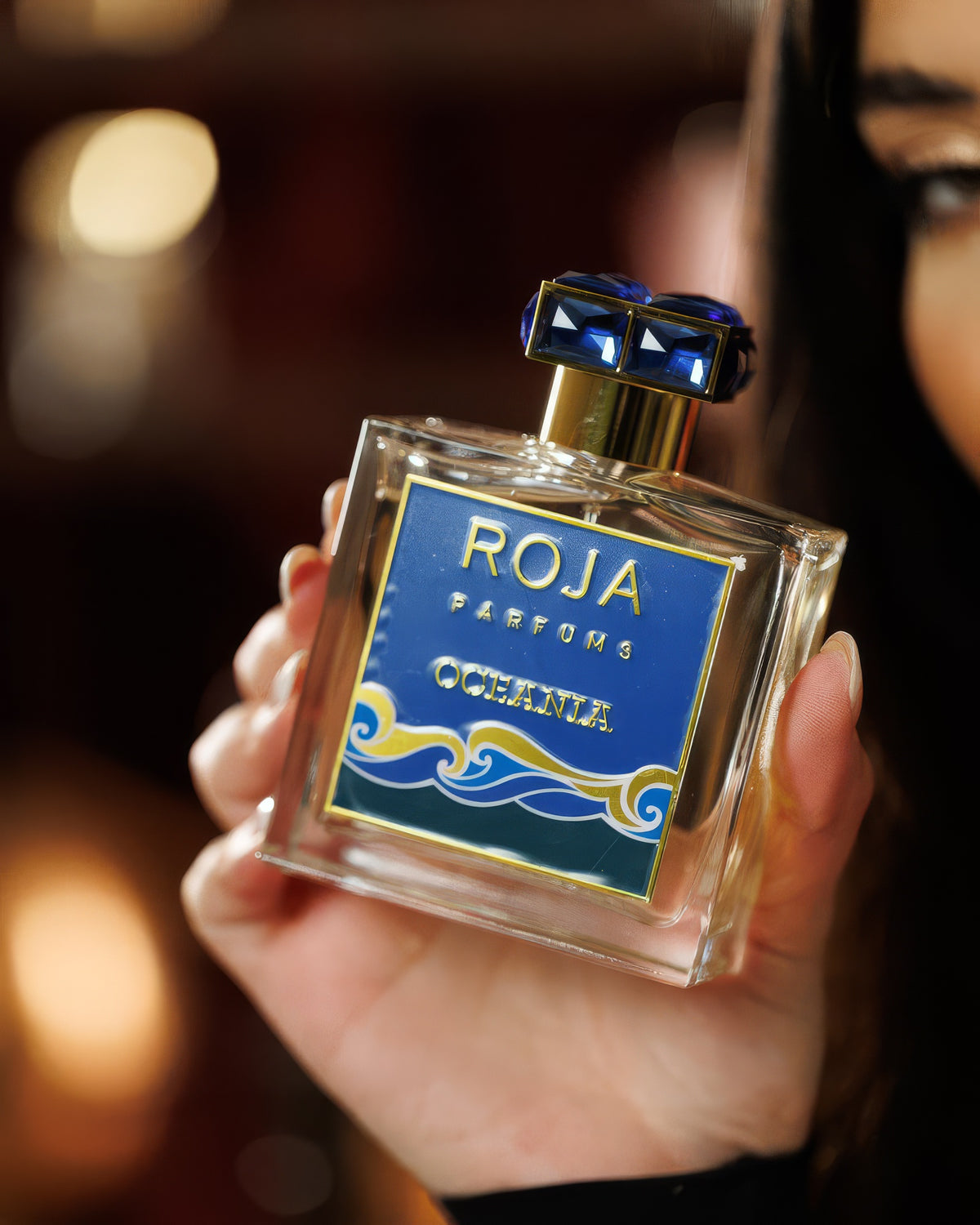 يد تمسك قارورة Roja Oceania أمام بوكيه ذهبي دافئ ووجه العارضة في الخلفية، عيّنة عطر مائي منعش