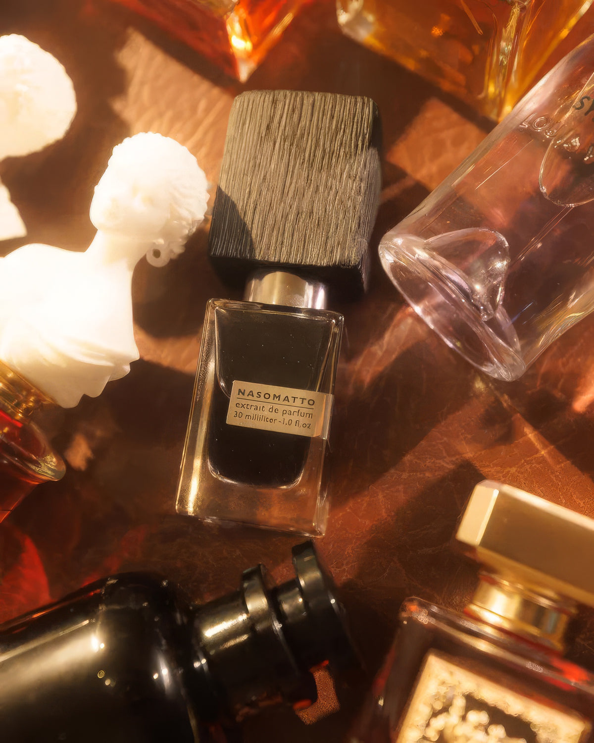 قارورة Nasomatto Black Afgano على الجلد بين العطور وتمثال نصفي فاتح، عيّنة عطر عود مدخّن