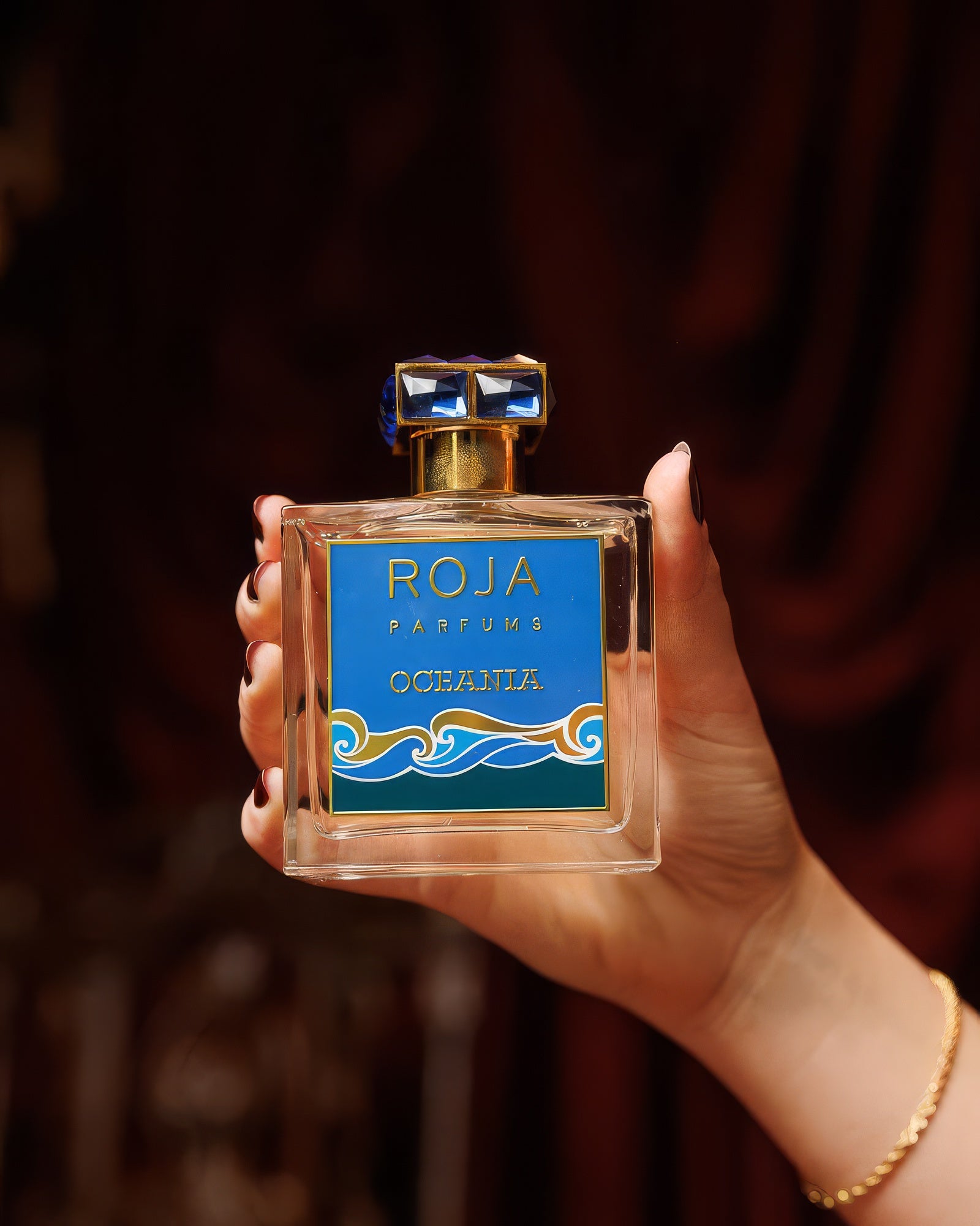 深紅のベルベットのドレープを背に Roja Oceania のボトルを掲げる手、アクアティックなエクストレ・ド・パルファムのサンプル
