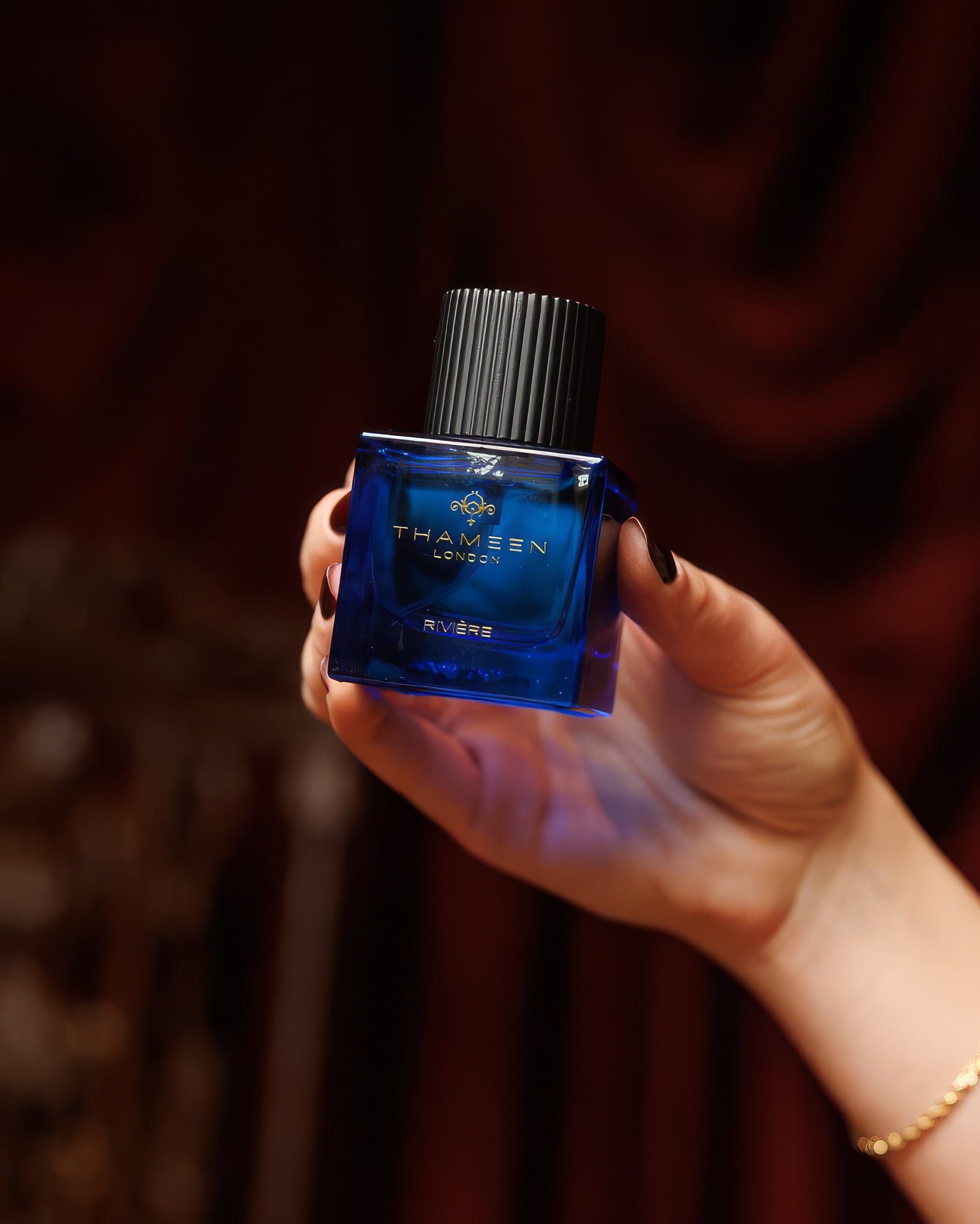 Hand with dark nails holding the Thameen Rivière bottle against deep red velvet drapery, spicy leather extrait de parfum sample