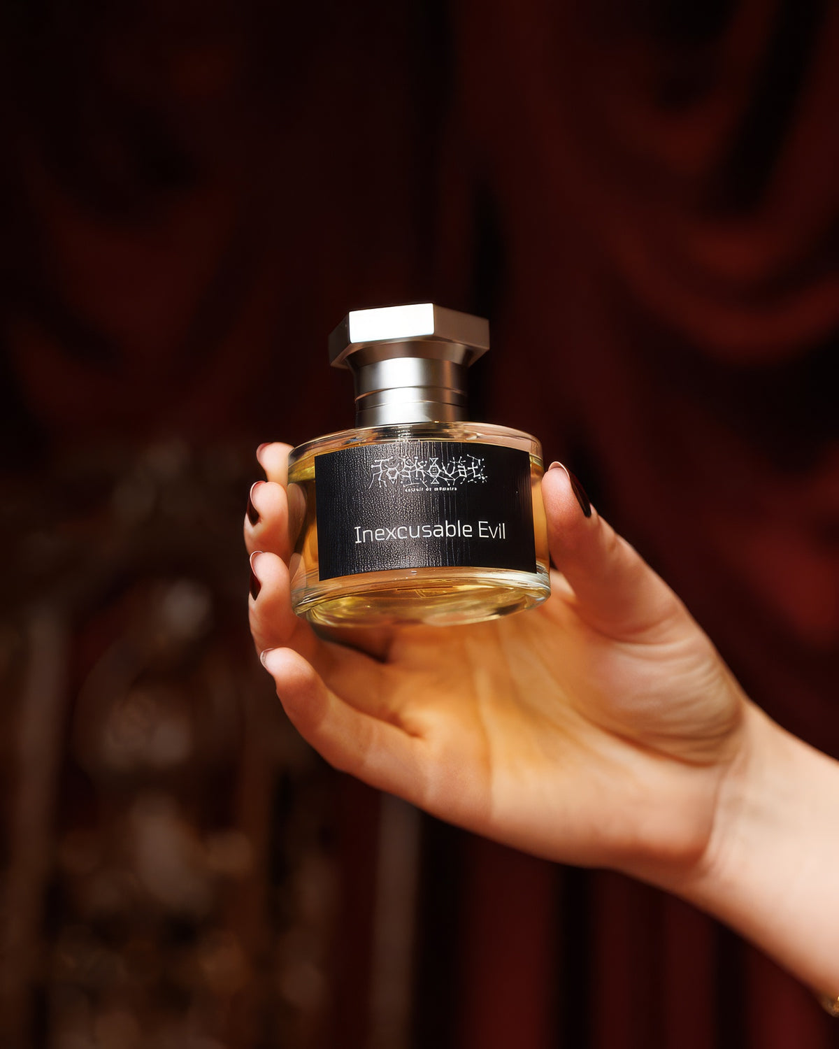 Mano che tiene il flacone Toskovat Inexcusable Evil davanti a velluto rosso intenso, campione di extrait de parfum scuro e fumoso