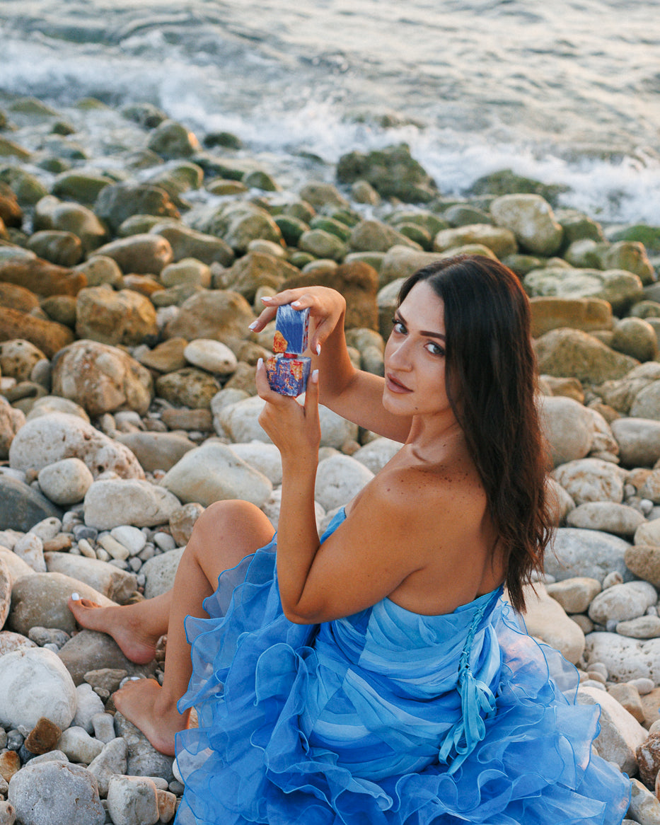 Adi Ale Van Hai Hui, flacone a cubo blu dipinto aperto da una donna in abito blu con volant su una spiaggia di ciottoli in riva al mare, decant extrait de parfum resinoso