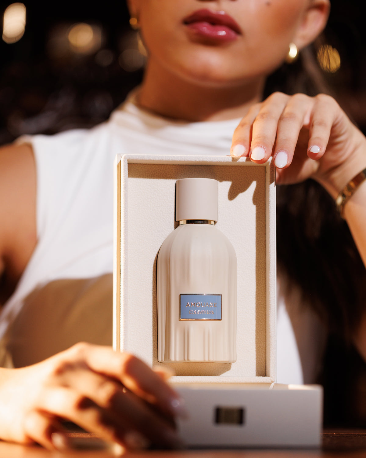 Amouage Elsewhere، قارورة كريمية مضلّعة بلوحتها الزرقاء قائمة في علبة عرض مفتوحة تحملها امرأة بالأبيض، وأضواء بار دافئة خلفها