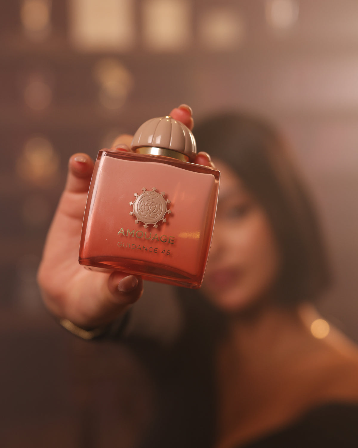 Amouage Guidance 46, bottiglia sfumata color pesca con tappo beige rigato porta verso l'obiettivo, donna sfocata dietro in un bokeh caldo, decant di fragranza floreale ambrata