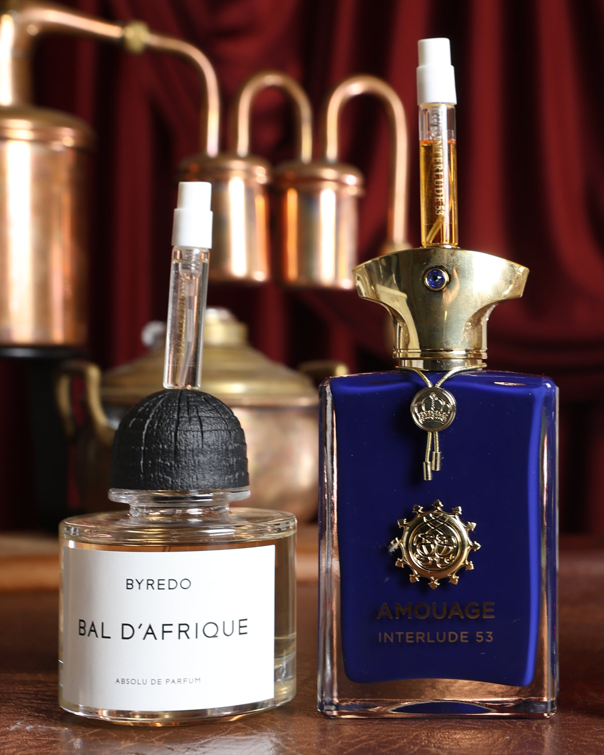 Altın kapağının üzerinde bir Gouttes Rares Interlude 53 şişeciği duran Amouage Interlude 53, yanında Byredo Bal d'Afrique Absolu, arkada bakır bir imbik, dumanlı tütsü notalı parfüm dekantı