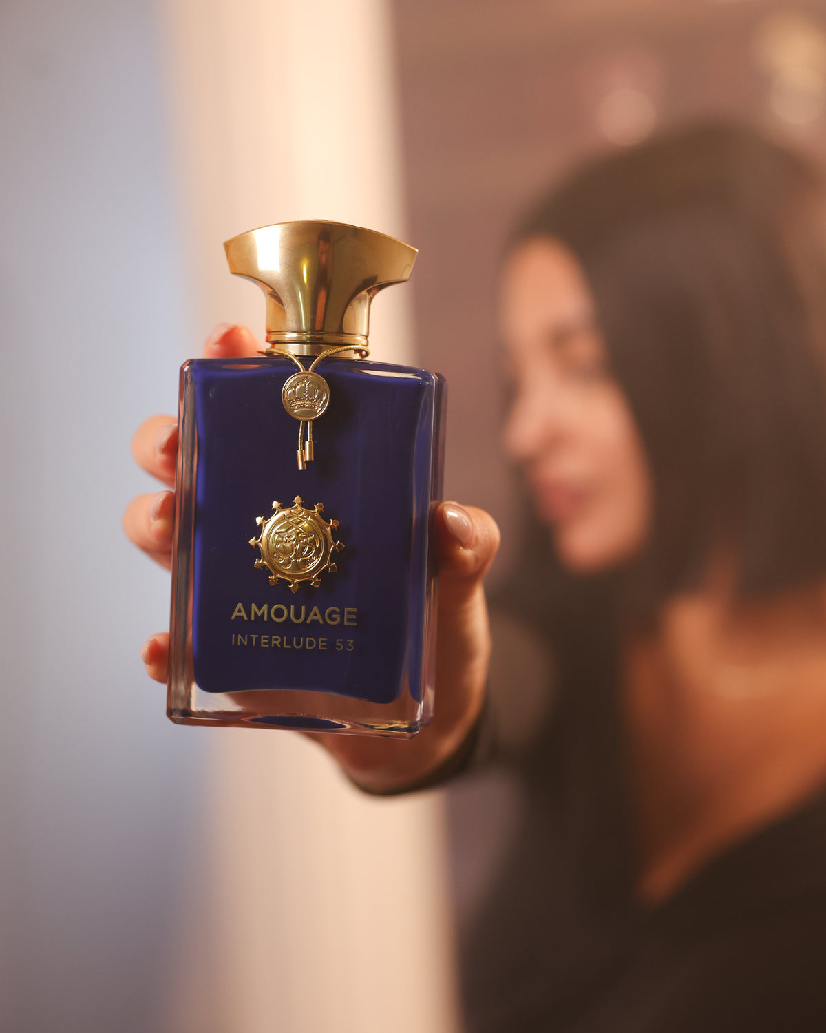 Amouage Interlude 53 altın kapaklı ve taç süslemeli mavi şişe kameraya doğru tutuluyor, arkada bulanık bir kadın, dumanlı tütsü extrait de parfum dekantı