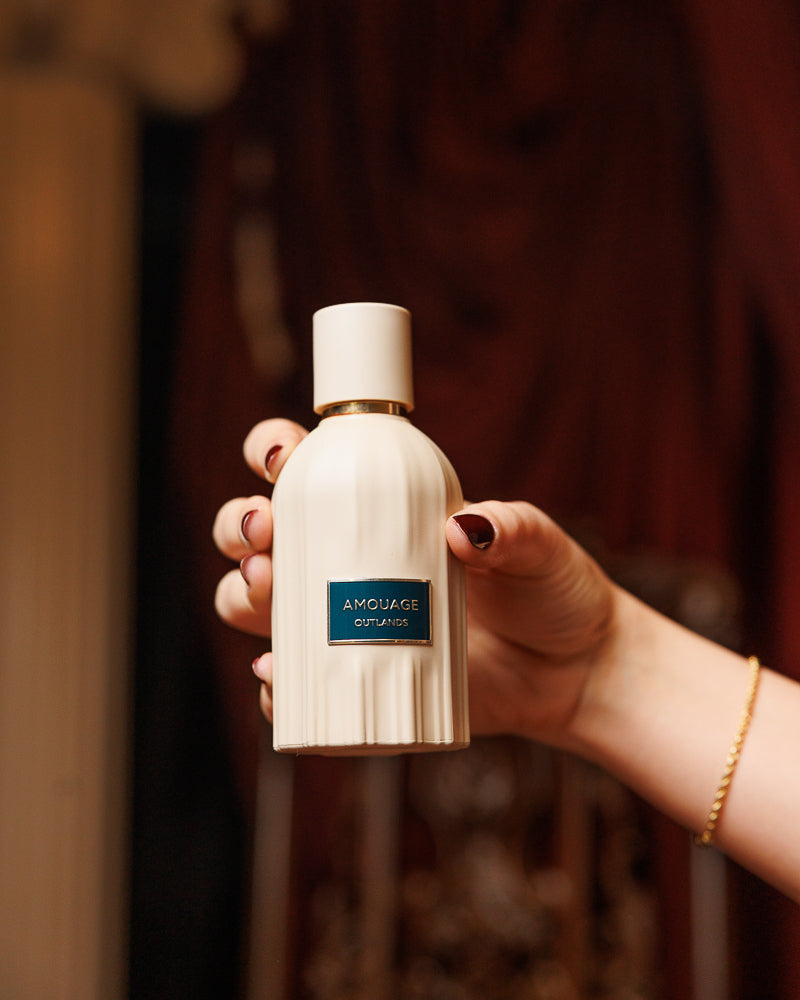 Flacon cannelé crème Amouage Outlands à l'étiquette bleu canard, tenu levé par une main aux ongles rouge foncé devant des tentures de velours rouge, décant de parfum de niche épicé et boisé