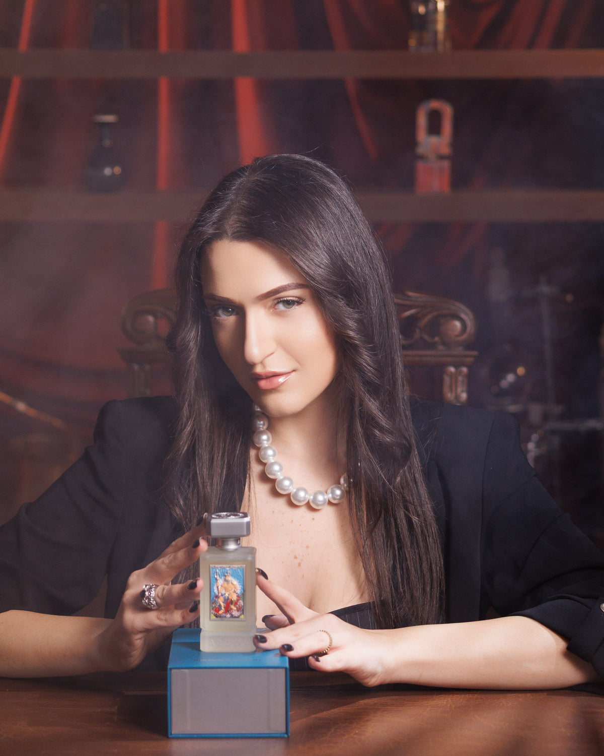 Argos Neptune's Trident frosted bottle on its blue box between a woman's hands at a wooden desk, black blazer and pearls, red curtain behind, citrus aromatic extrait decant