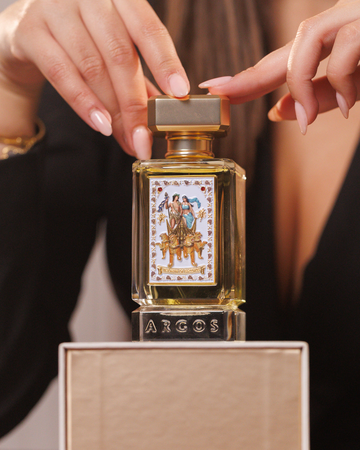 Argos Triumph of Bacchus Extrait krem rengi kutusunun üzerinde, parmak uçları beyaz emaye panelin üstündeki altın kapakta, likörlü amber extrait de parfum numunesi