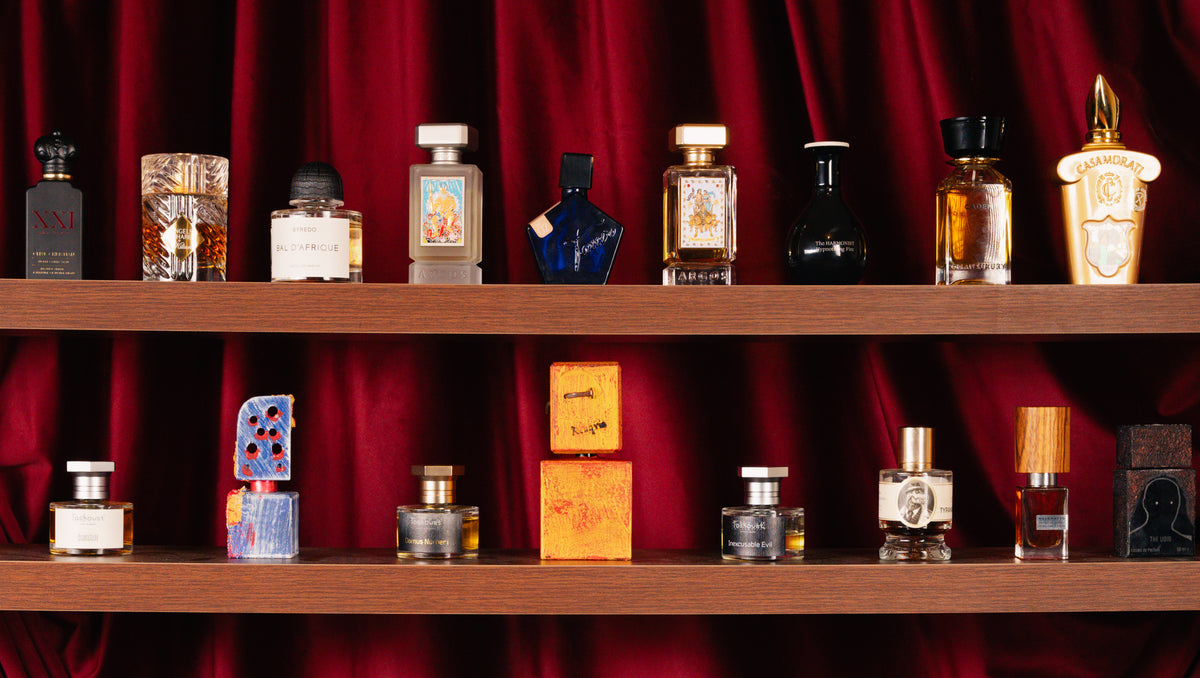 Byredo Bal D'Afrique Absolu, Argos Neptune's Trident, Kilian Paris Angels’ Share, Clive Christian Blonde Amber and Toskovat bottles on two wooden shelves before red velvet