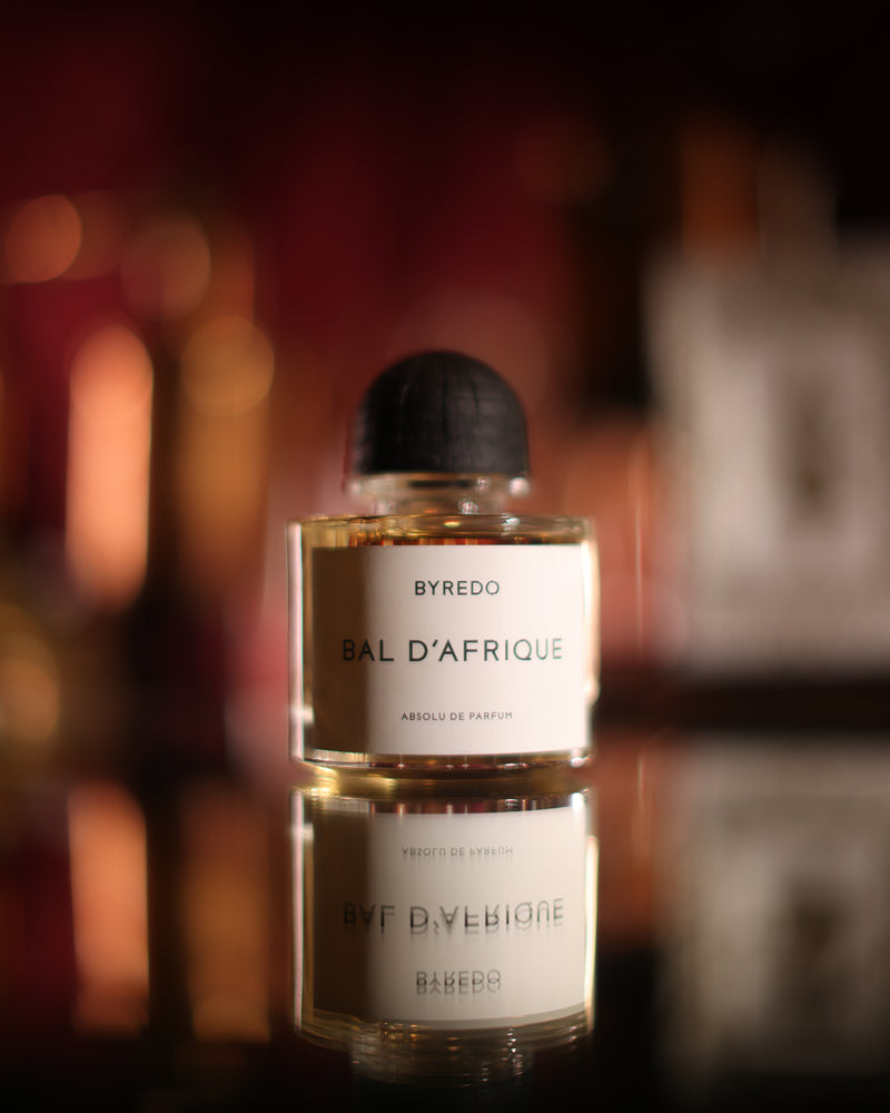 Byredo Bal D'Afrique Absolu, the clear flacon with its charred wood cap reflected in a black mirror, red bottle shelves blurred behind