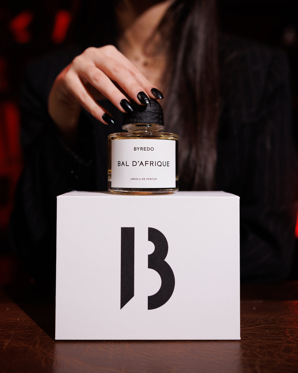Byredo Bal D'Afrique Absolu on its white B box, a hand with black nails resting on the black textured cap, woman in a dark blazer blurred behind, woody niche perfume decant