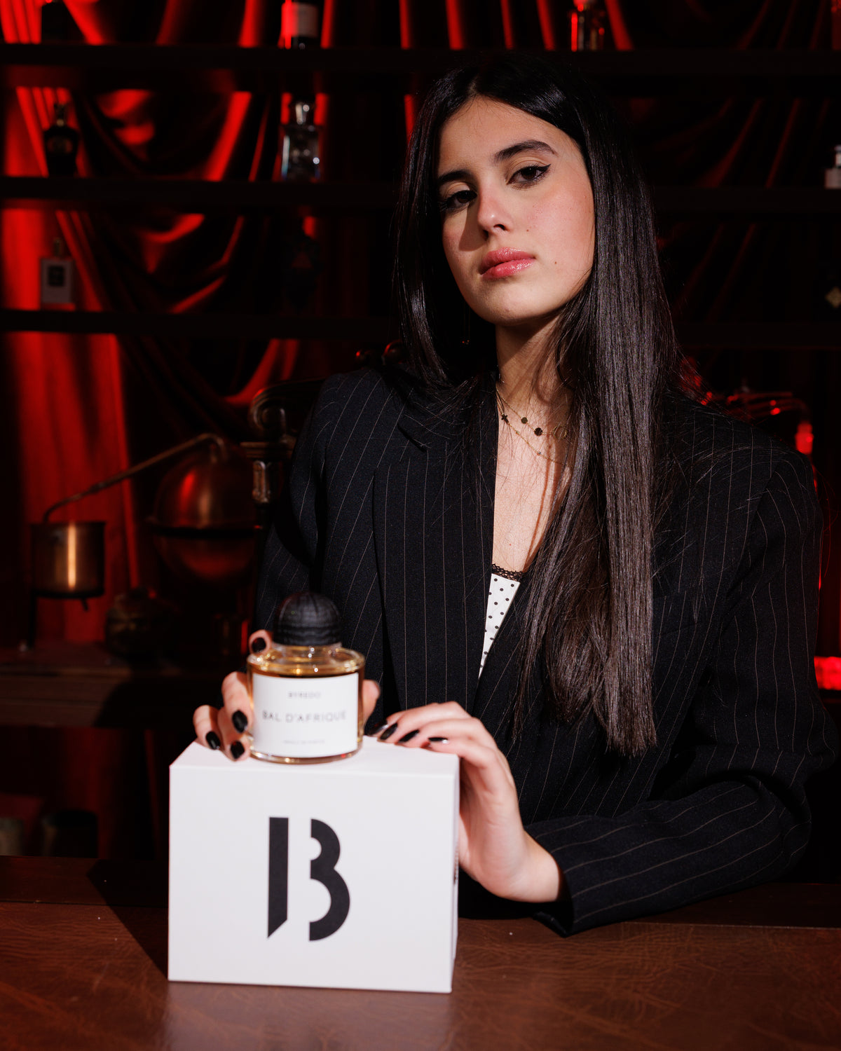 Byredo Bal D'Afrique Absolu on its white B box, a woman in a pinstripe blazer with her hands around the bottle, red velvet curtain and shelves behind, woody niche perfume decant