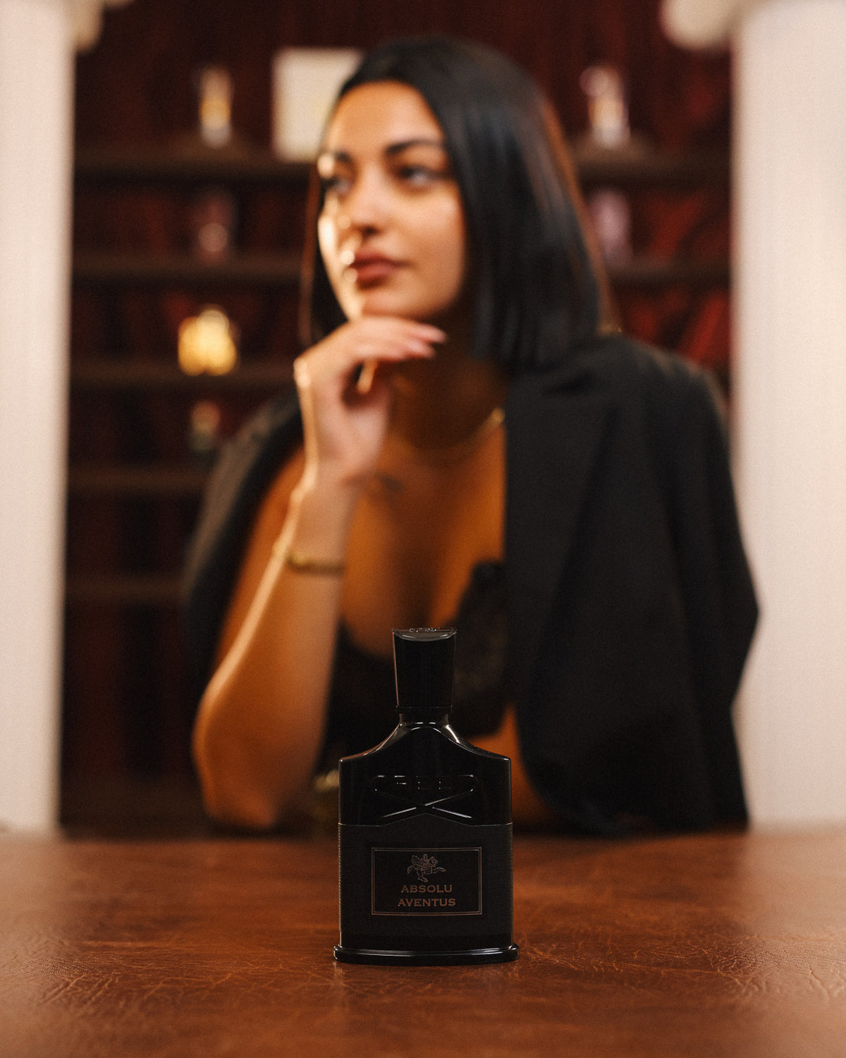 Creed Absolu Aventus, frasco negro solo sobre una mesa de cuero, mujer con blazer negro apoyando el mentón en la mano detrás, decant de perfume nicho amaderado frutal