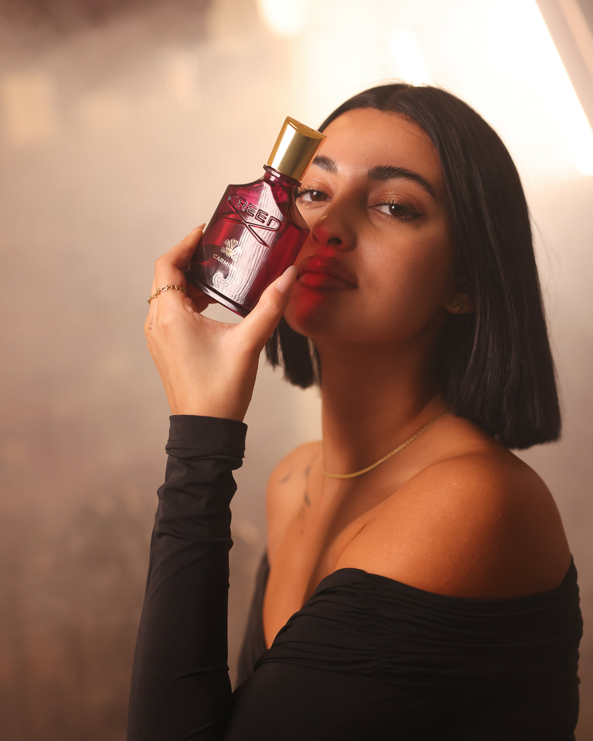 Creed Carmina bottle pressed to the cheek of a woman with a sleek bob in an off-shoulder black top, warm smoky haze, spicy floral fragrance decant