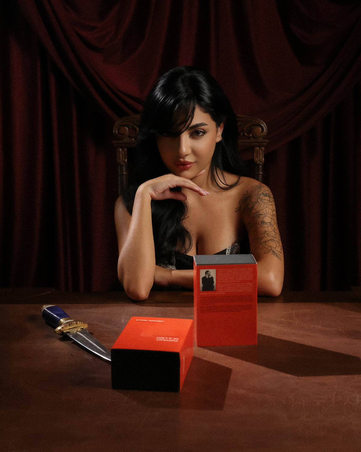 Frédéric Malle Portrait of a Lady, dos cajas rojo anaranjado y una daga sobre una mesa ante una mujer y cortinas de terciopelo rojo, decant de perfume nicho de rosa e incienso