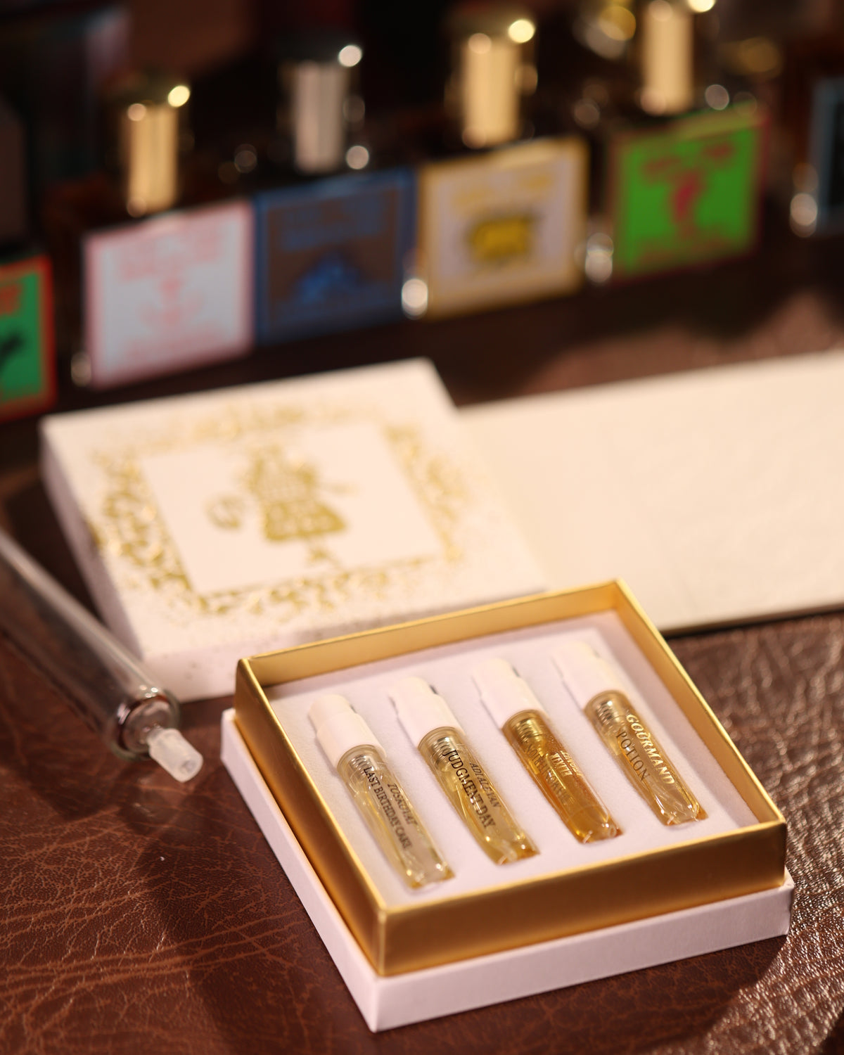Gouttes Rares Niche Perfume Discovery Sets 4 x 2ml مفتوحة على جلد بني، أربعة بخّاخات بسعة 2ml في الصينية الذهبية، غطاء برقائق ذهبية في الخلفية، حقنة زجاجية على اليسار، علبة عيّنات منتقاة بعناية