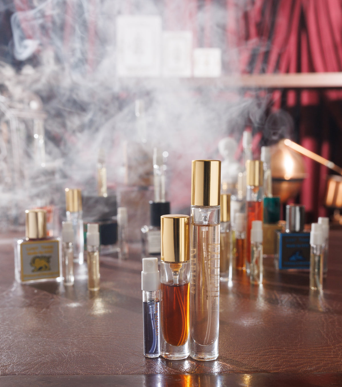 Gouttes Rares decants, a Black Afgano 2 ml sample, a 5 ml atomiser and a Delina Exclusif 10 ml, on a leather desk in fog before a red curtain, niche perfume decant