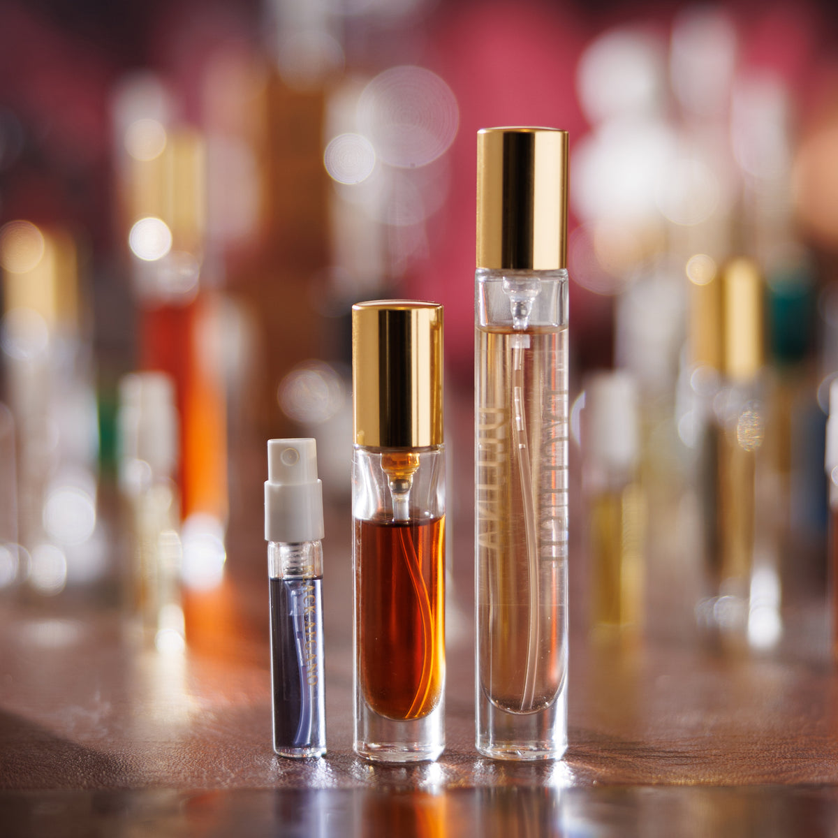 Decant Gouttes Rares in tre formati su una scrivania lucida: un campione da 2 ml di Black Afgano, un atomizzatore ambrato da 5 ml e un atomizzatore da 10 ml di Delina Exclusif, bokeh rosso sullo sfondo