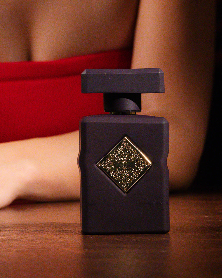 Initio Side Effect, frasco ameixa fosco com emblema dourado em losango sobre uma mesa de madeira diante de uma mulher de blusa vermelha, decant de perfume nicho de tabaco licoroso