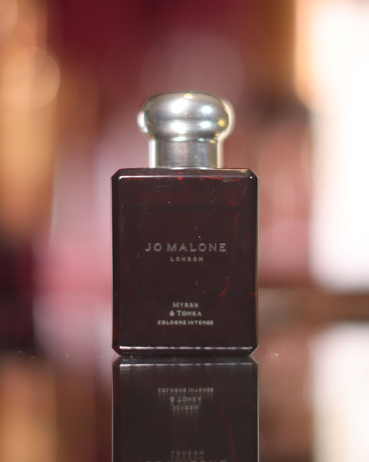 Dunkelbraune Jo Malone Myrrh & Tonka Cologne Intense Flasche mit Silberkappe, gespiegelt auf einem schwarzen Spiegel vor unscharfen roten und bernsteinfarbenen Lichtern, Abfüllung eines Amber-Gourmand-Nischenparfums