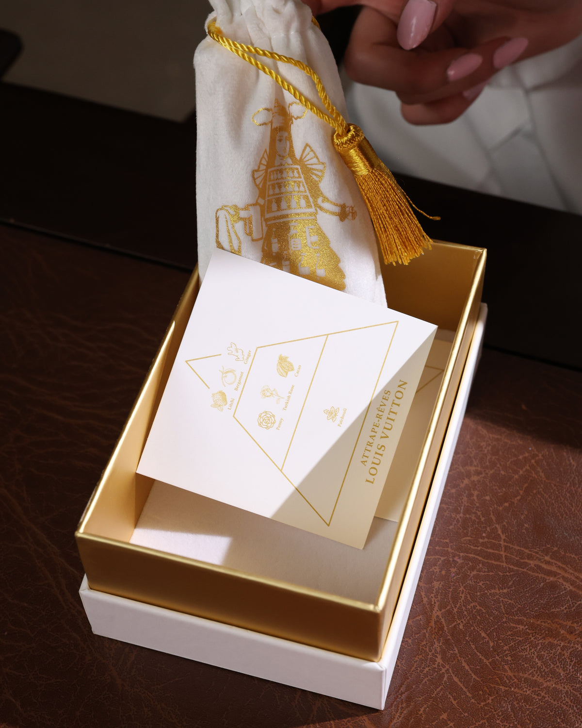 Duftnotenkarte von Louis Vuitton Attrape-Rêves mit Litschi, Pfingstrose, türkischer Rose und Patchouli in einer goldgefütterten Box von Gouttes Rares, eine Hand hebt den Beutel an, Nischenparfum-Abfüllung