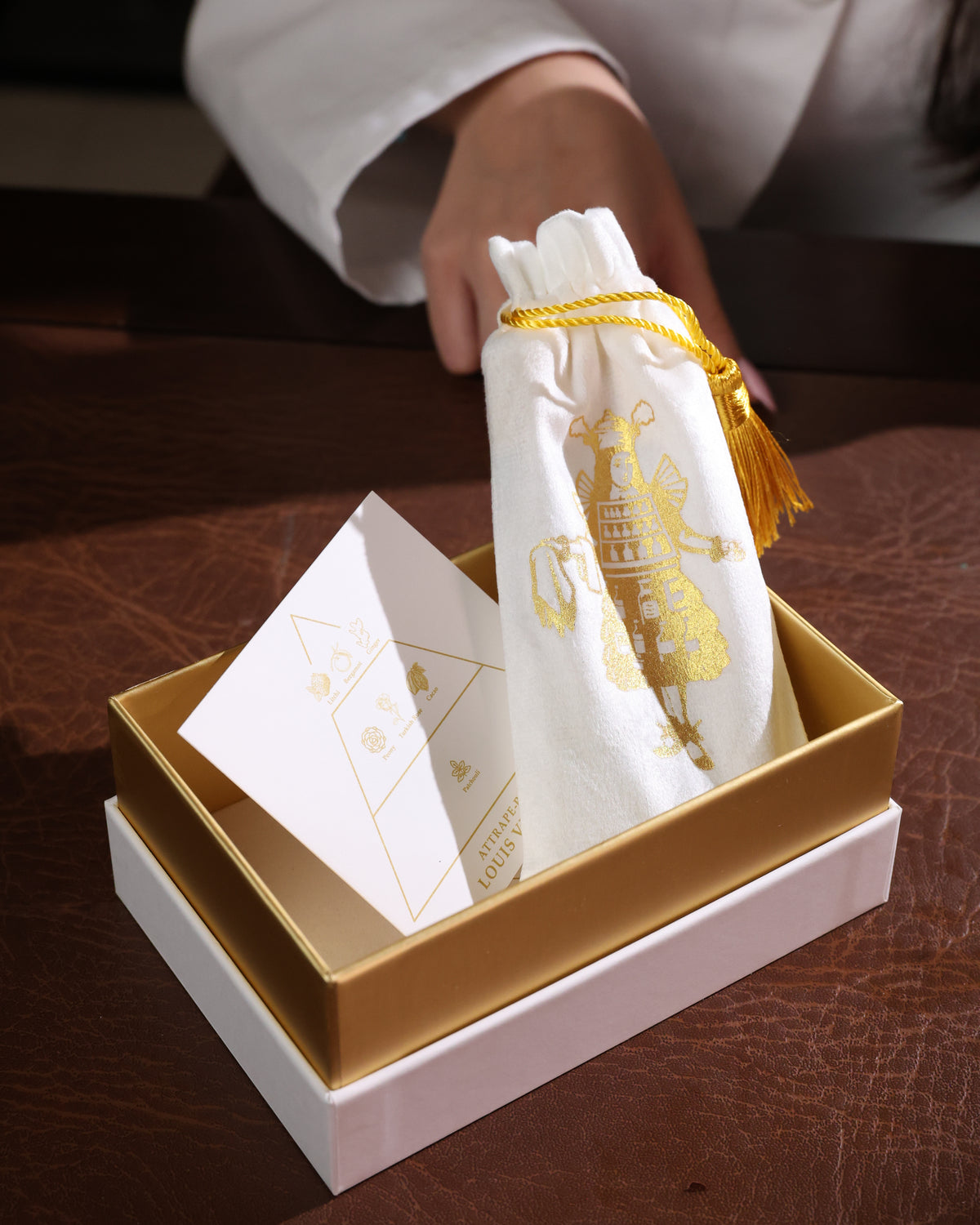 Schachtel von Gouttes Rares mit goldenem Tablett, weißem Samtbeutel mit goldener Quaste und der Duftnotenkarte zu Louis Vuitton Attrape Rêves auf einem Lederschreibtisch, Abfüllung eines fruchtig-floralen Dufts