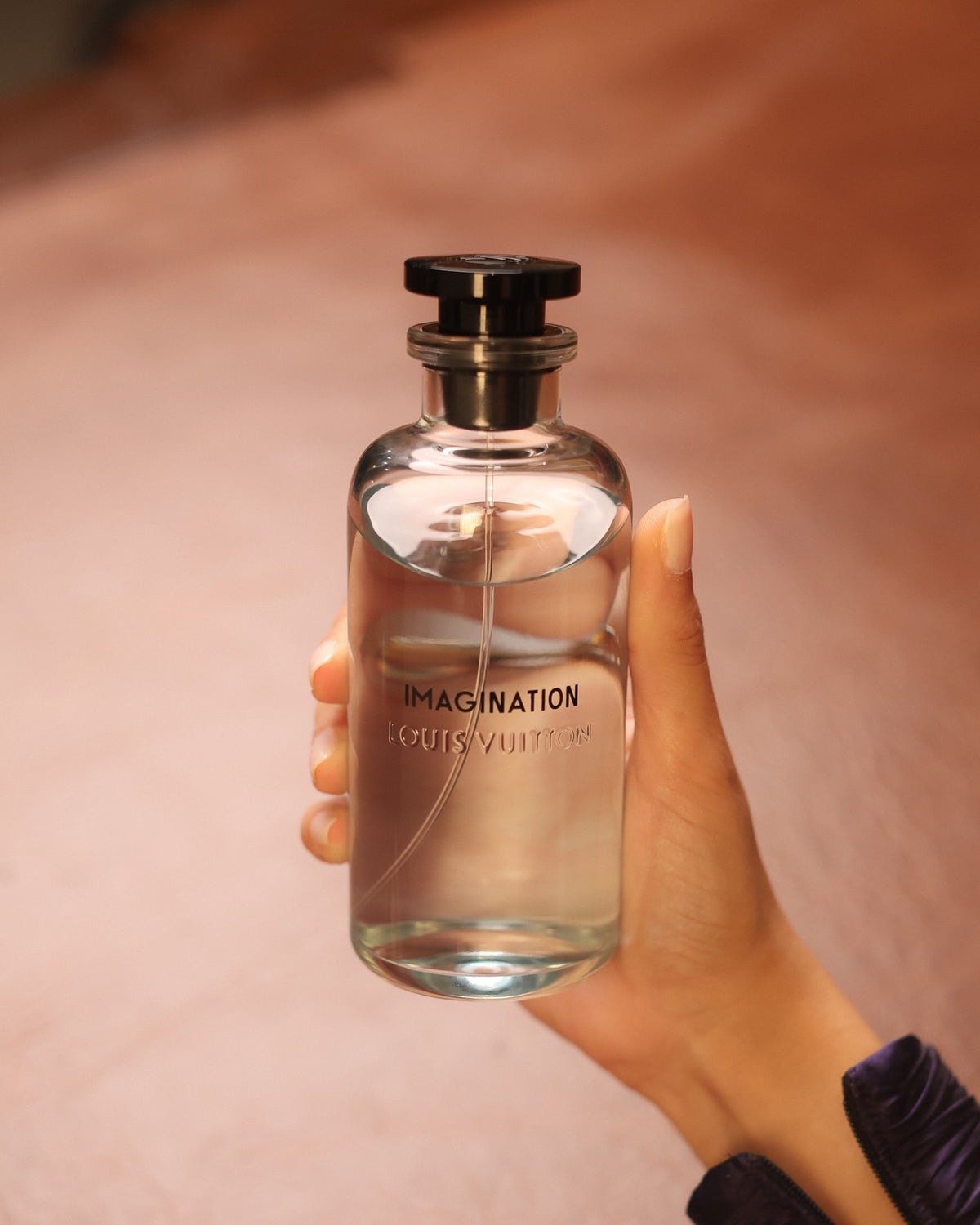 Louis Vuitton Imagination 더스티 핑크 블러를 배경으로 한 손에 곧게 든 검은 캡의 투명 보틀, 프레시한 시트러스 니치 향수 디캔트