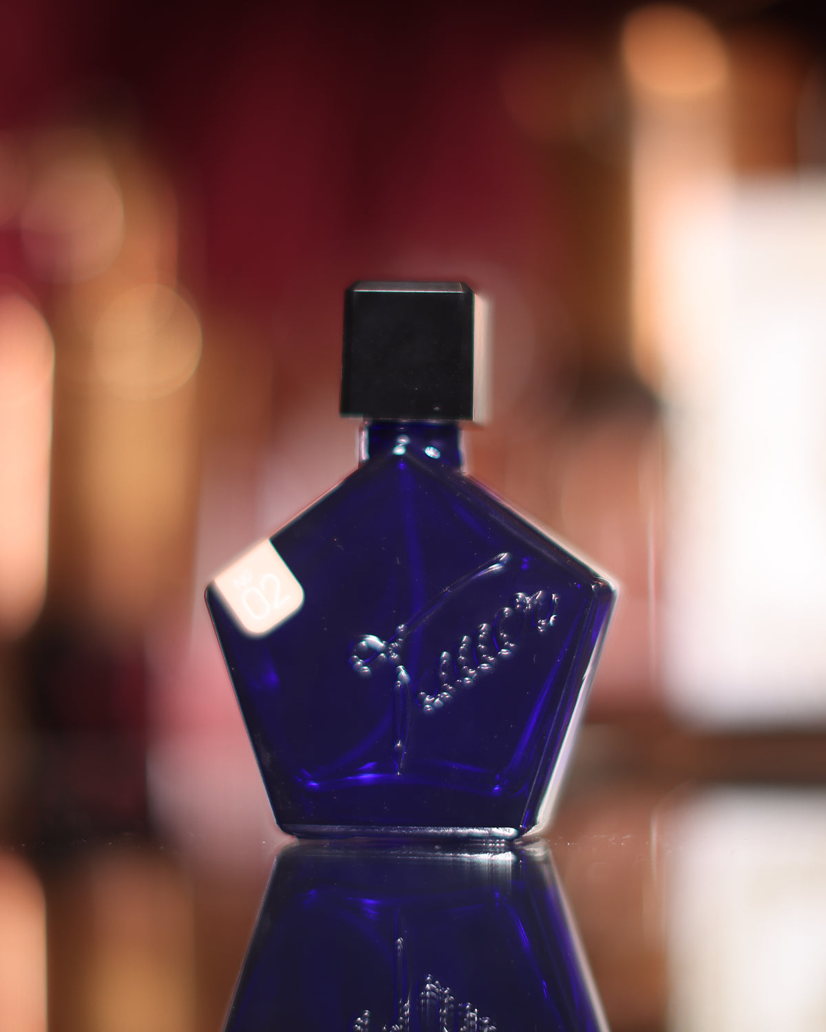 Bottiglia pentagonale cobalto Tauer Perfumes L’Air du Désert Marocain con tappo nero e targhetta 02 su uno specchio nero, caldo bokeh rosso sullo sfondo, decant di profumo di nicchia ambrato resinoso