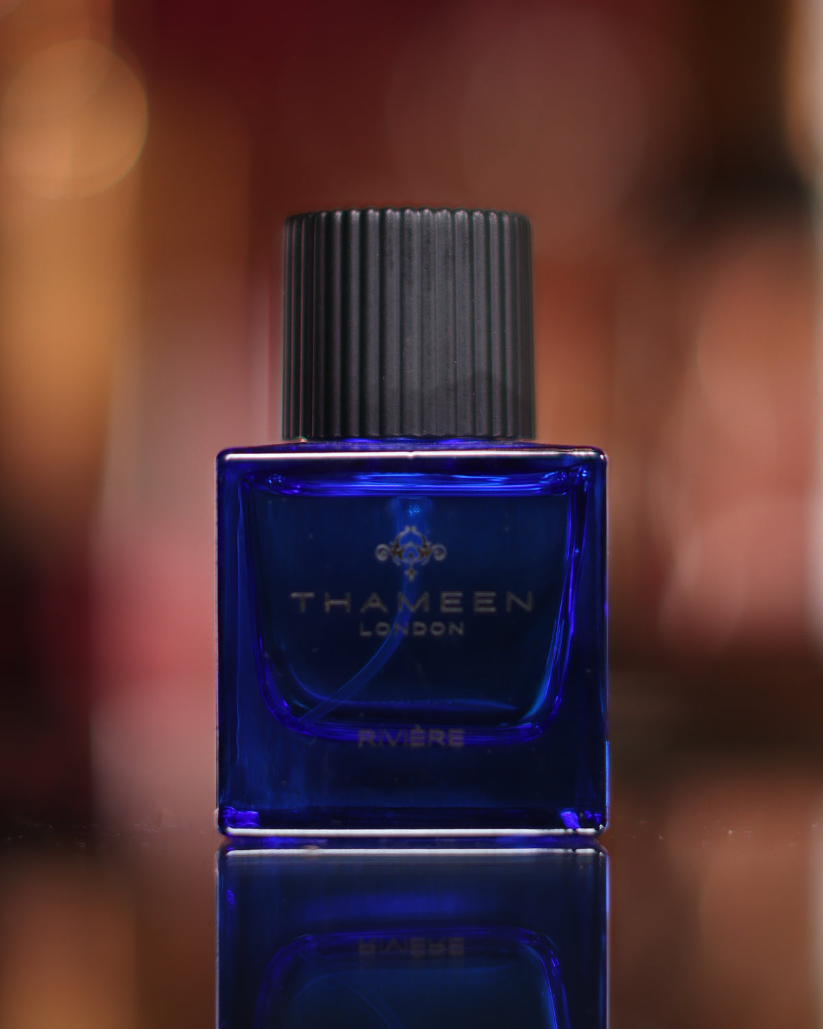 Kobaltblaue würfelförmige Thameen Rivière Flasche mit gerippter schwarzer Kappe, gespiegelt auf einem schwarzen Spiegel vor warmen unscharfen Lichtern, Abfüllung eines Leder-Rosen-Extrait de Parfum